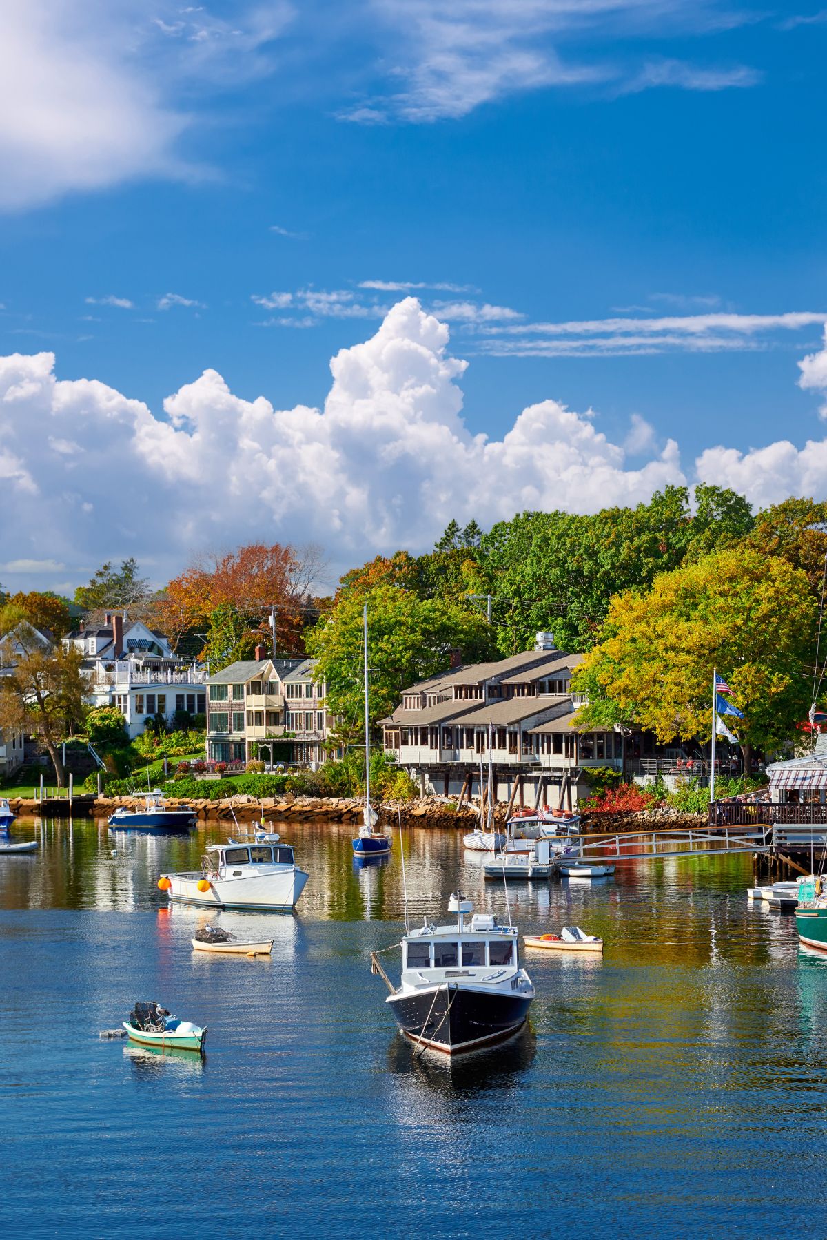 Boats floating in the harbor in Ogunquit, Maine, with colorful houses and trees lining the shoreline in the background.