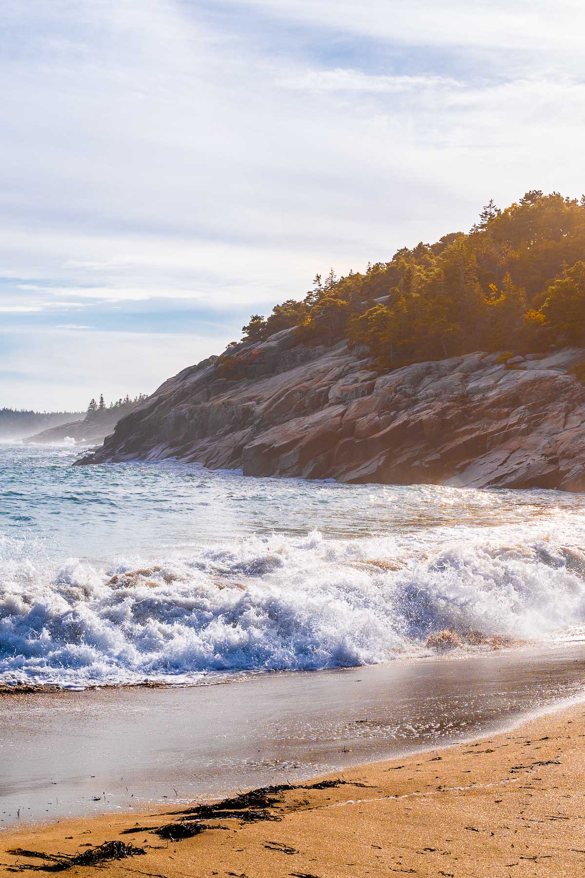 Waves crashing onto the sandy shore at Sand Beach in Acadia National Park, with rocky cliffs and pine trees in the background.