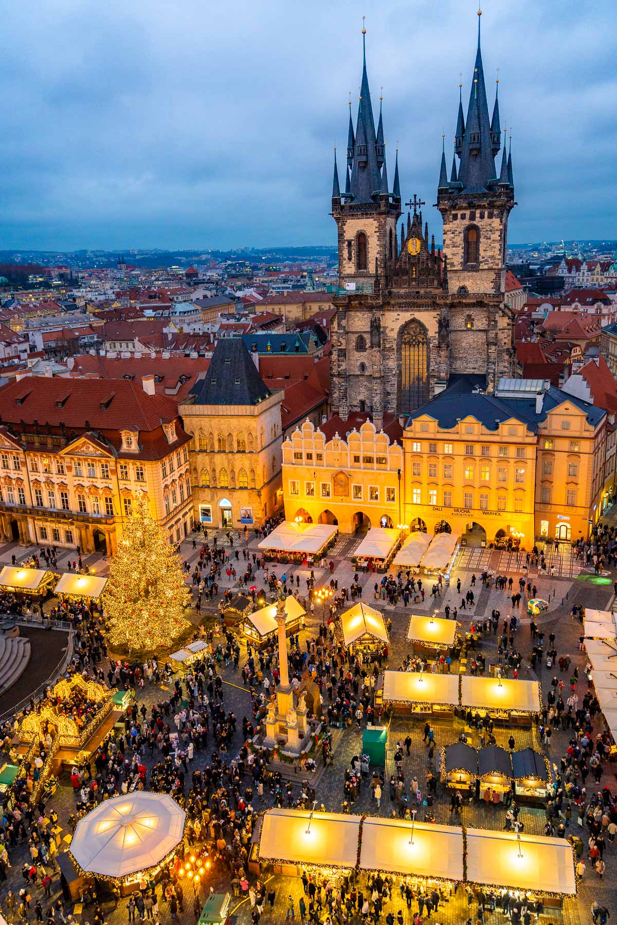 Aerial view of the Christmas market in Old Town Square, Prague, showing glowing stalls, a huge lit Christmas tree, and the twin spires of Týn Church rising behind a row of pastel buildings.