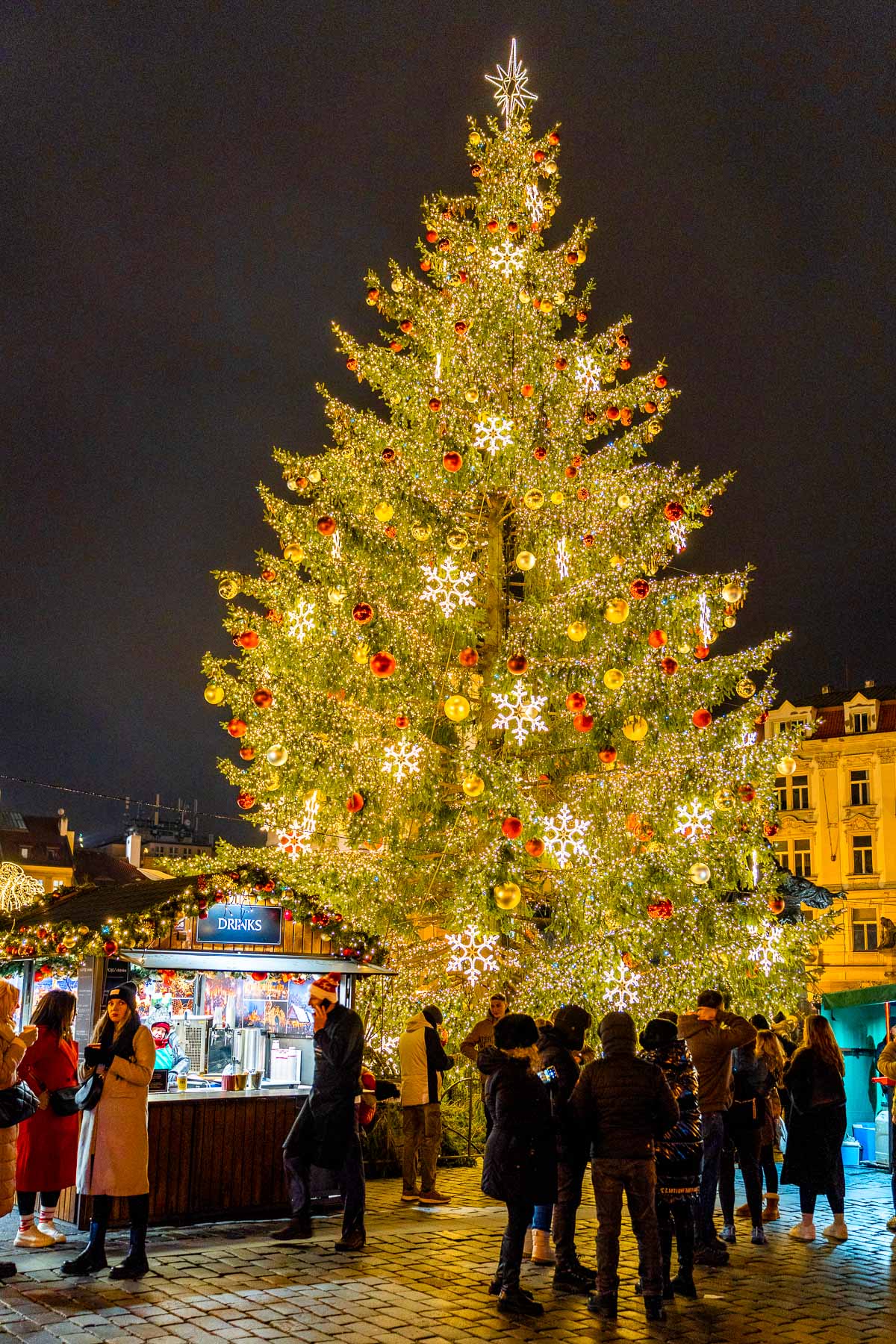 People standing near a drink stall next to a tall Christmas tree covered in golden lights and red ornaments at Prague’s Old Town Square during the evening.