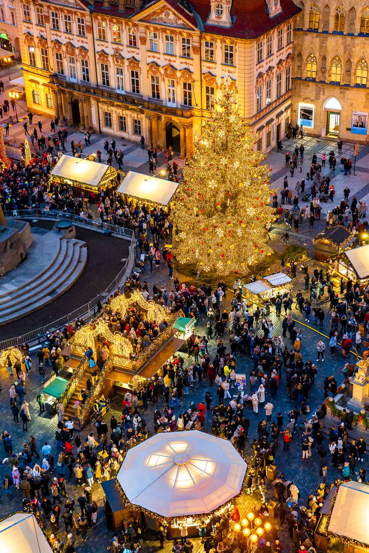 View from above of Prague’s Old Town Square during the Christmas market, with a large lit-up tree in the center, rows of decorated market stalls, and a crowd of people walking around.