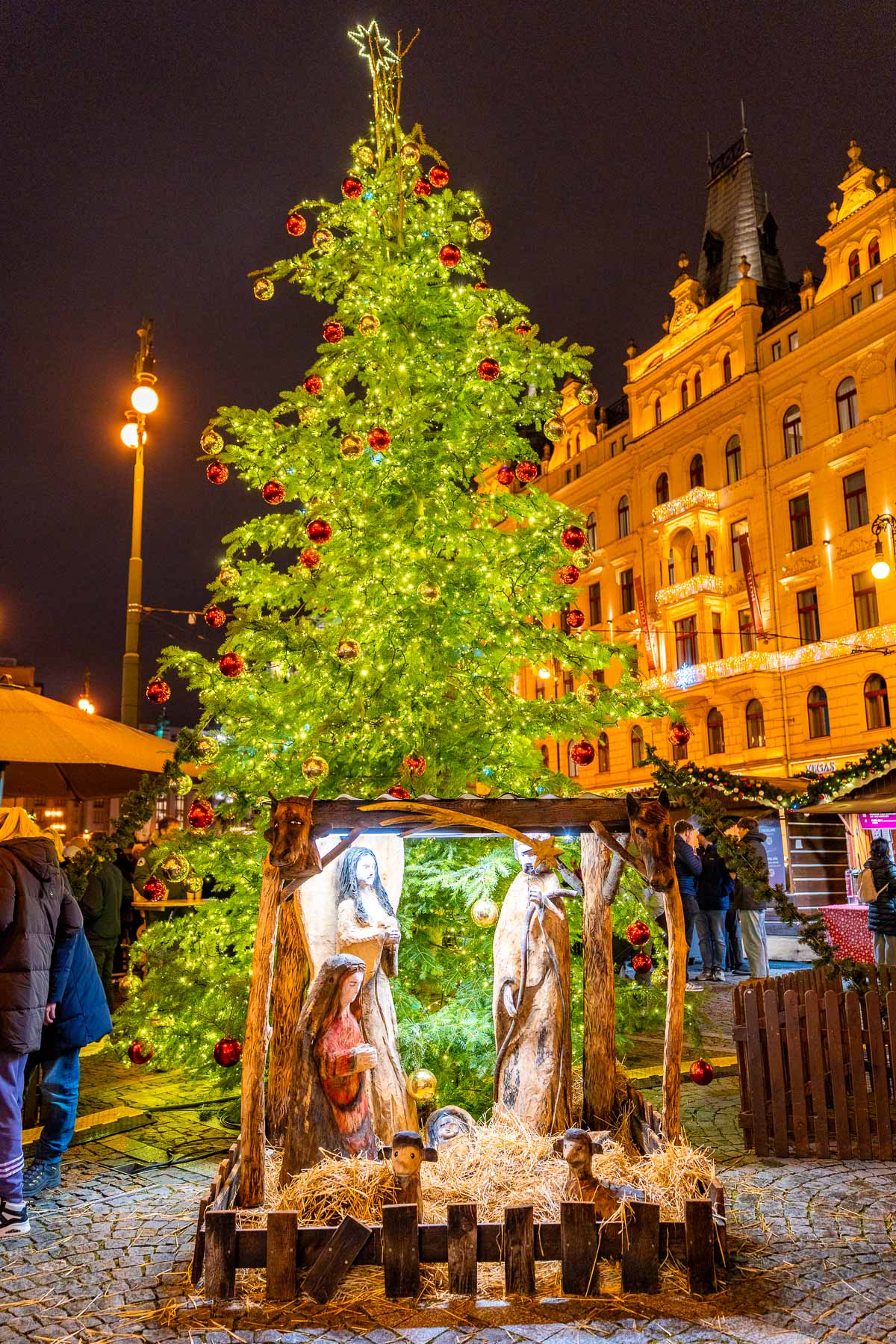 A Christmas tree decorated with red and gold ornaments stands behind a wooden nativity scene at the Christmas Market on Republic Square in Prague.