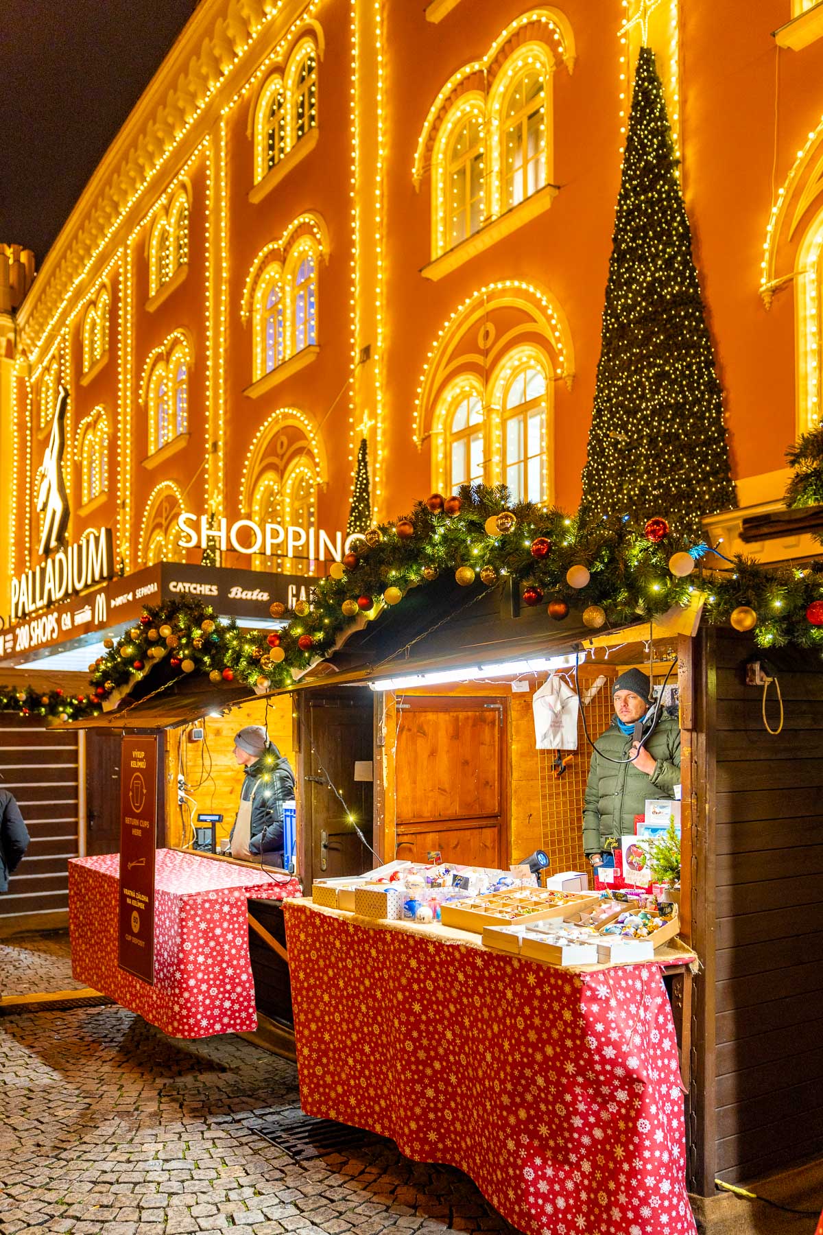 Market stalls decorated with garlands and lights line the Christmas Market on Republic Square in Prague, with the Palladium shopping center lit up in the background.