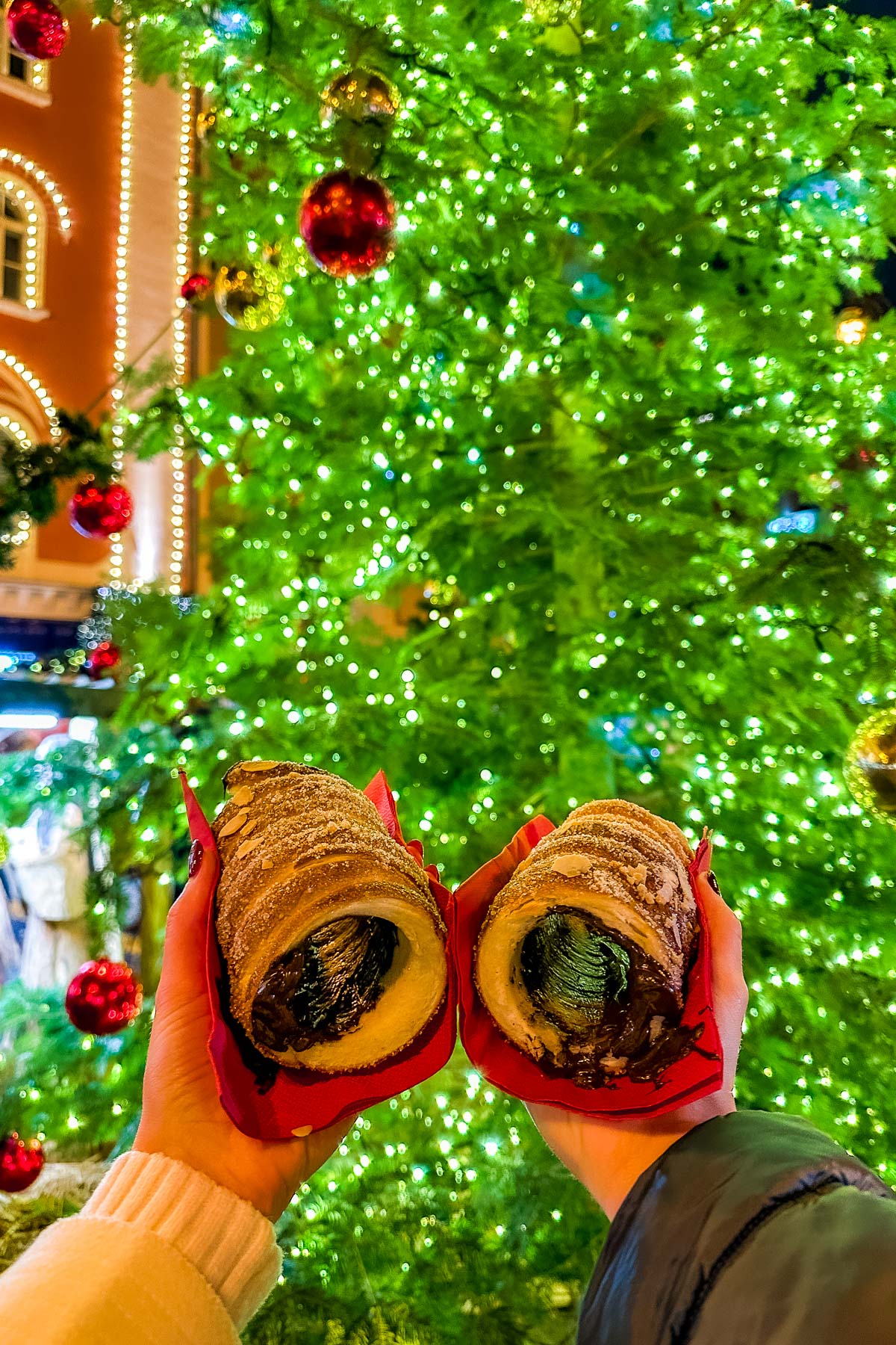 Two trdelníks filled with chocolate are held in front of a large Christmas tree at the Christmas Market on Republic Square in Prague.