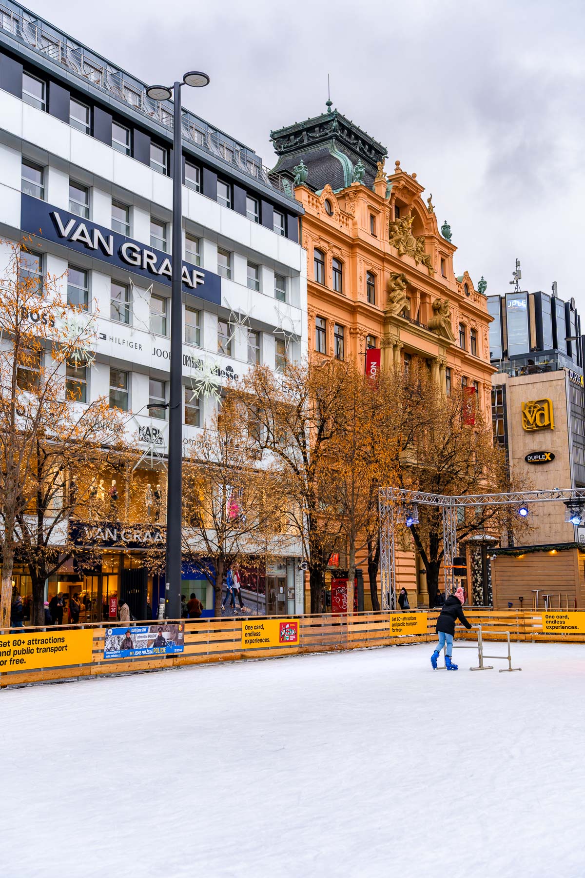 An ice-skating rink set up in Wenceslas Square, Prague, with a child skating in the foreground and a backdrop of shops and historic buildings.