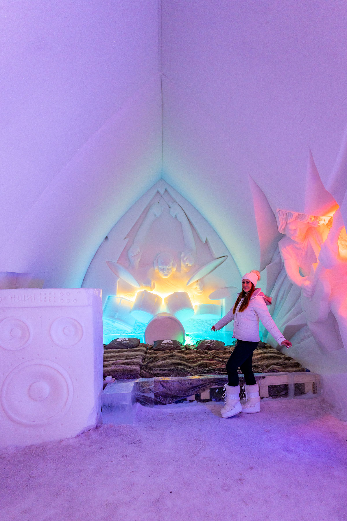A woman inside the Arctic SnowHotel in Lapland, with a colorful ice sculpture wall and a bed made of snow and ice, lit by glowing lights.