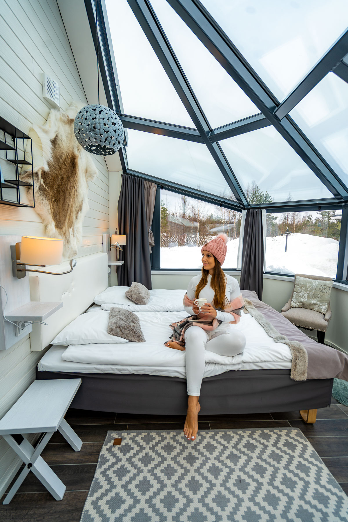 A cozy glass igloo room at Santa's Igloos Arctic Circle in Lapland, with a woman sitting on the bed under the large glass ceiling, snowy scenery visible outside.