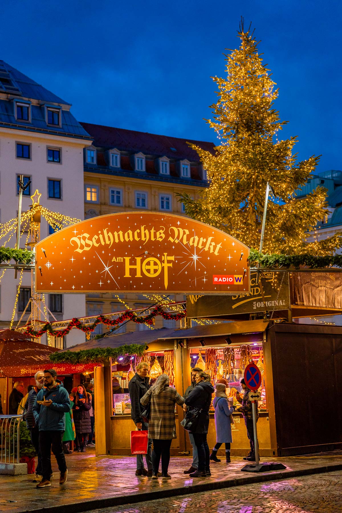 People browsing food stalls at the Am Hof Christmas market in Vienna, with festive lights and a large decorated Christmas tree in the background.
