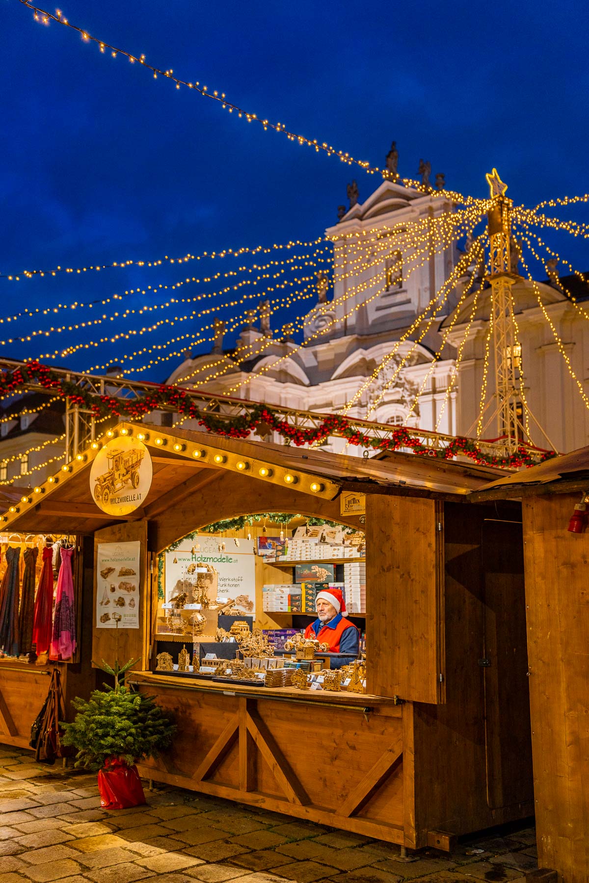 A vendor wearing a Santa hat selling handcrafted wooden models at the Am Hof Christmas market in Vienna, surrounded by glowing string lights.