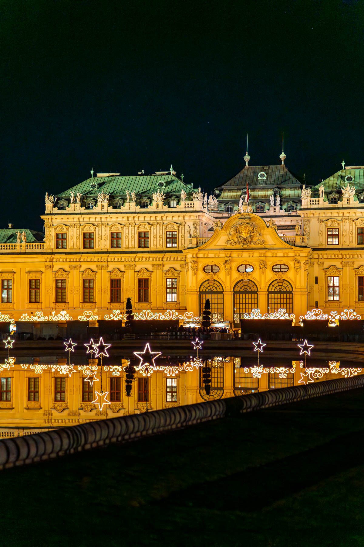 The Belvedere Palace in Vienna beautifully lit at night, with glowing star-shaped lights reflecting in the water in front of the Christmas market.