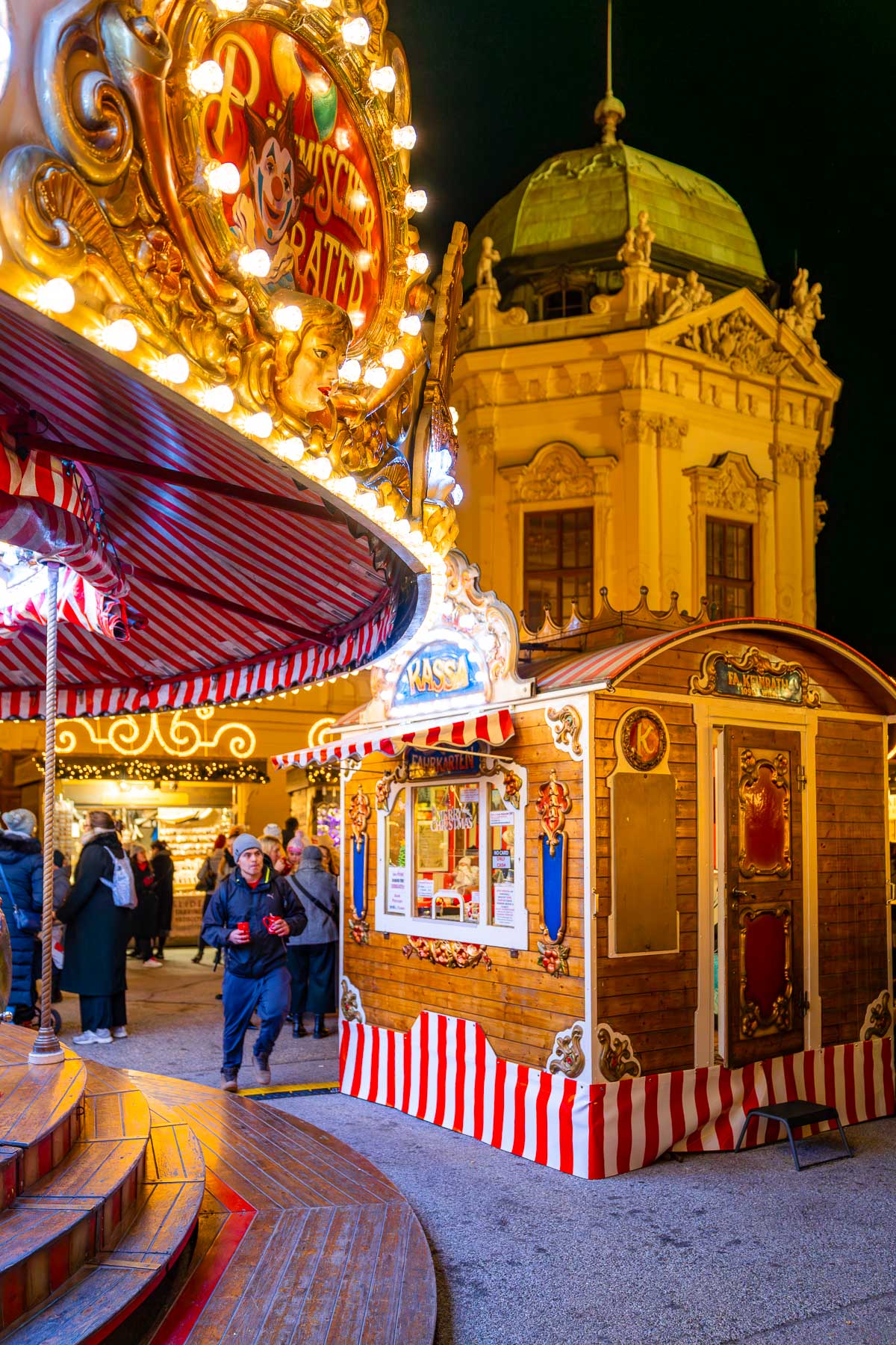 A carousel and wooden ticket booth at the Belvedere Palace Christmas market in Vienna, surrounded by twinkling lights and baroque architecture.