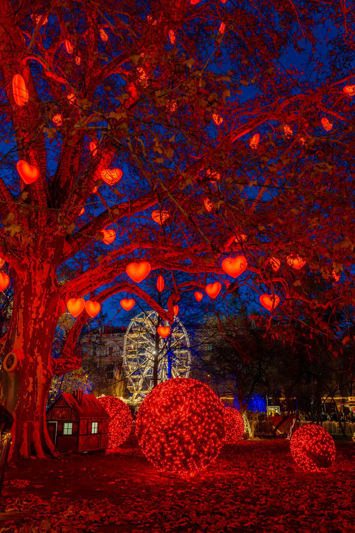 Trees decorated with glowing red heart-shaped lights and illuminated spheres at the Rathausplatz Christmas market in Vienna, with a Ferris wheel in the background.