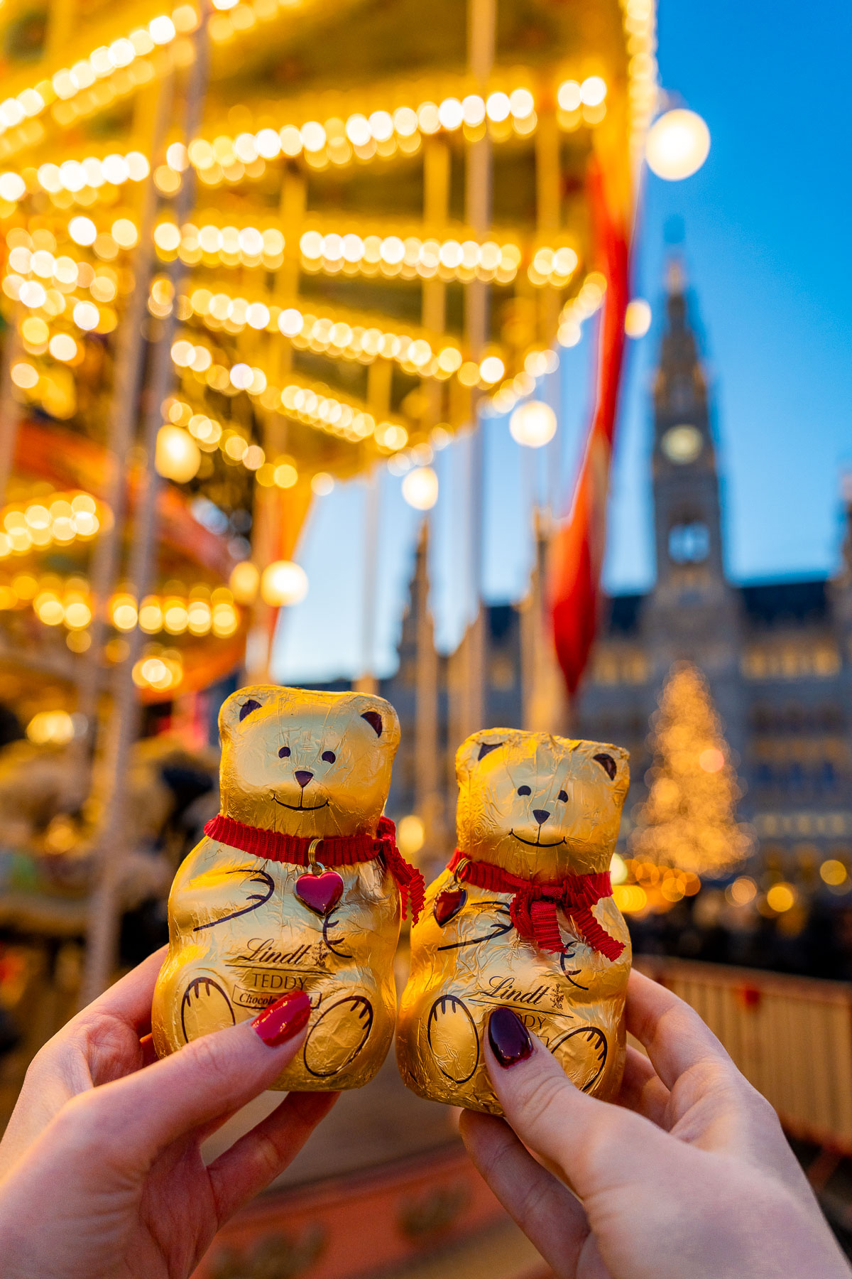 Two Lindt chocolate bears held up in front of the glowing carousel lights at the Rathausplatz Christmas market in Vienna.