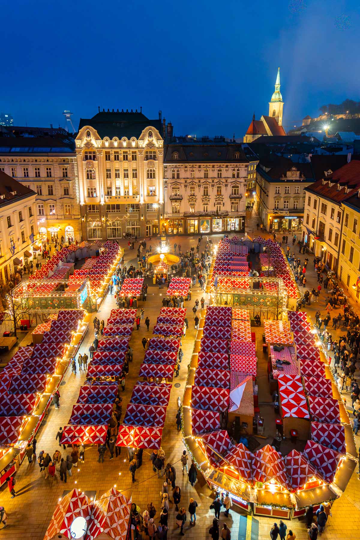 View of the Hlavné Square Christmas Market in Bratislava at night, with rows of red and white roofed stalls, string lights, crowds filling the square, and surrounding historic buildings lit up.