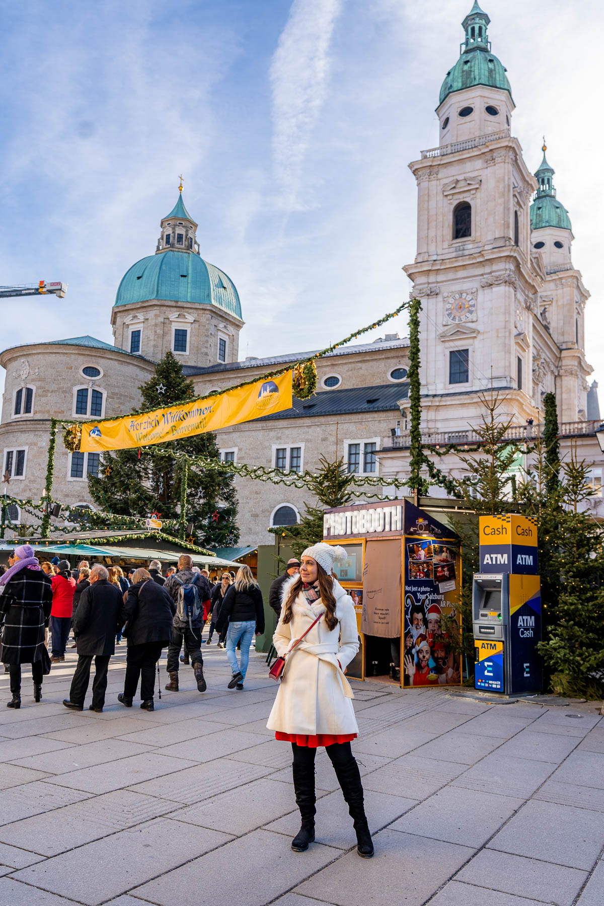A woman standing in front of Salzburg Cathedral at the Residenzplatz Christmas Market in Salzburg, wearing a white coat, red dress, and knit hat, with decorated stalls, Christmas trees, and crowds behind her.