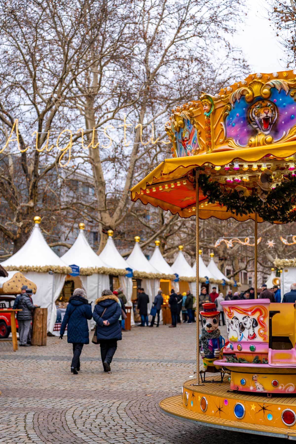 A colorful carousel and rows of white market tents at the Augustusmarkt in Dresden, with people strolling along the cobblestone street.