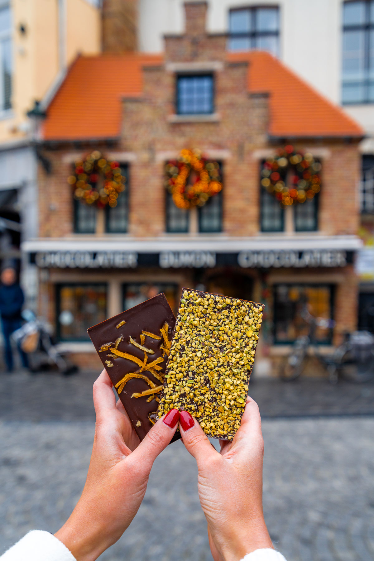 Two hands holding chocolate bars topped with candied orange and nuts in front of the Chocolatier Dumon shop in Bruges, decorated with festive wreaths.