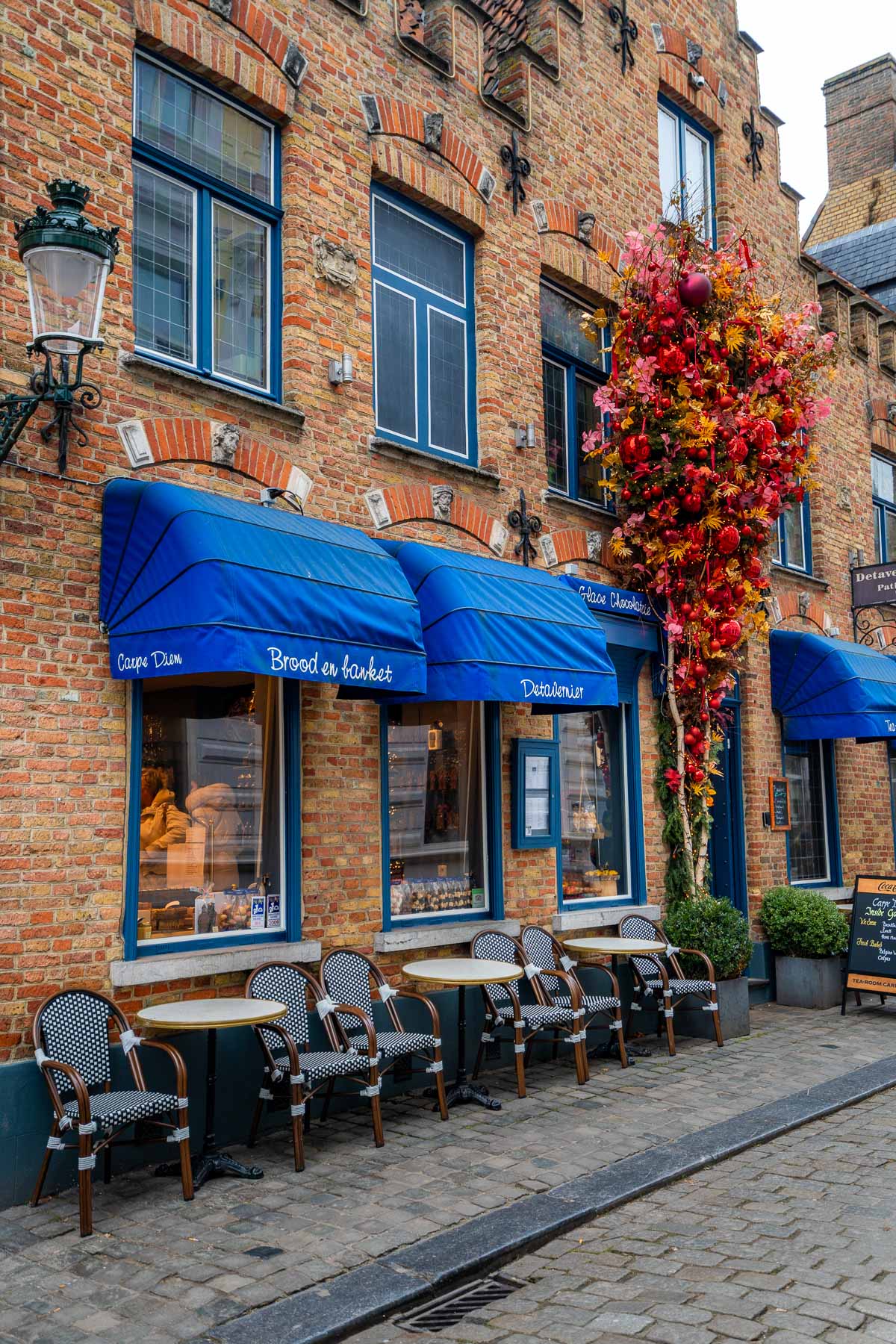Cozy café in Bruges with blue awnings, outdoor seating, and a tall arrangement of red and gold holiday ornaments by the entrance.