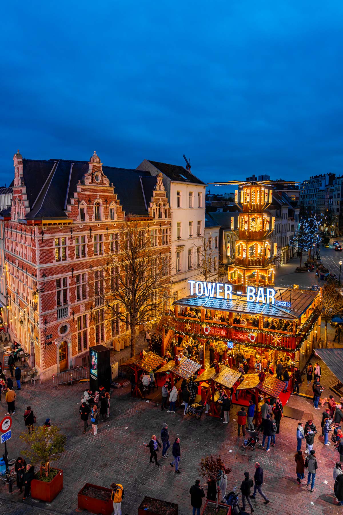 Evening view of the Tower Bar and wooden stalls at the Christmas market in Marché aux Poissons, Brussels, glowing with festive lights against the blue sky.