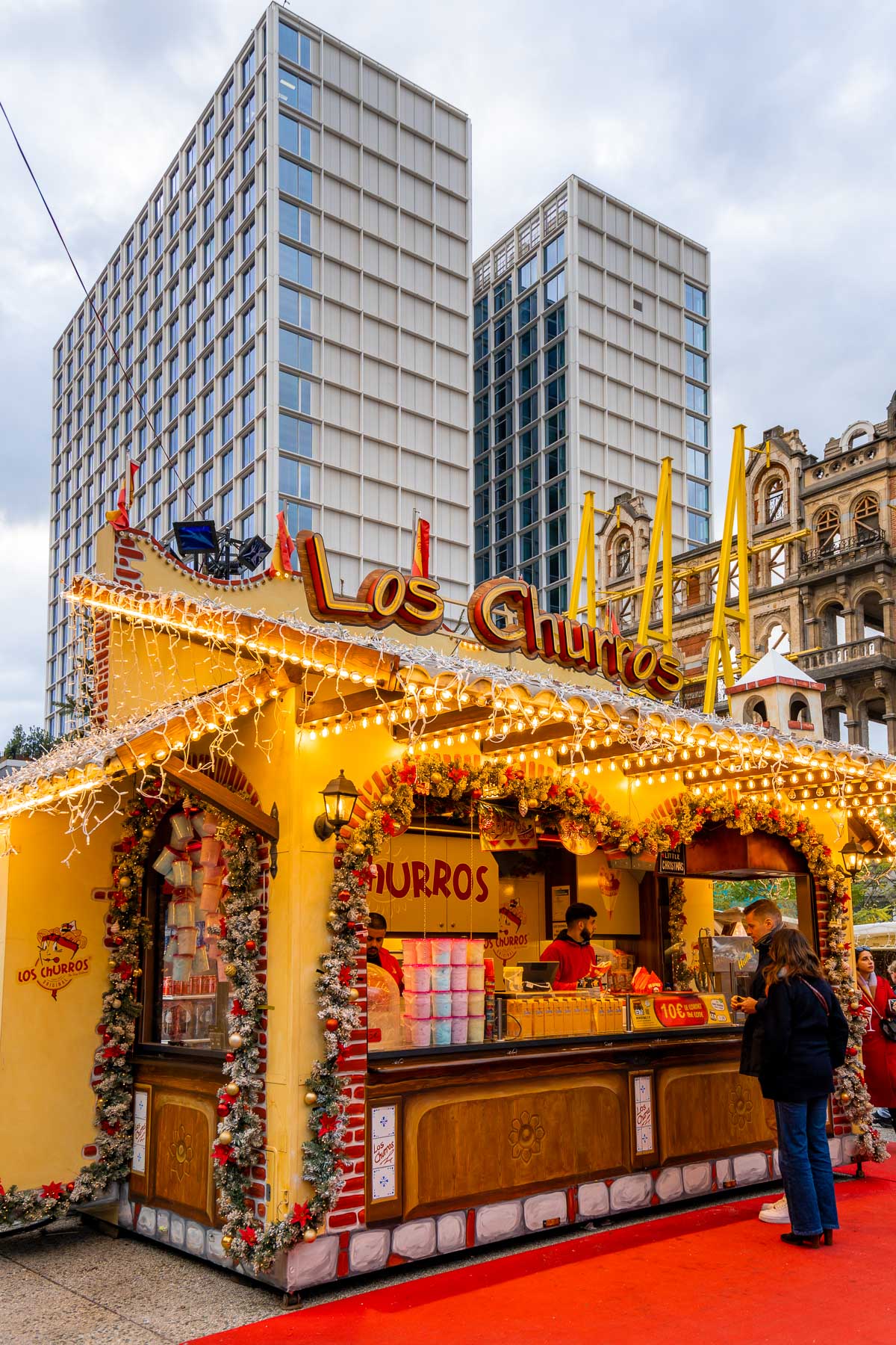 Los Churros food stall decorated with garlands and string lights at the Brussels Christmas market, set against modern high-rise buildings.