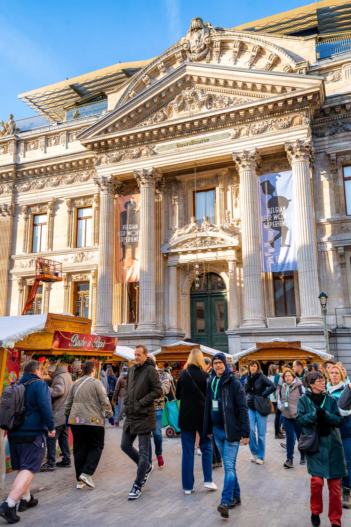 Daytime scene at the Christmas market in front of the Brussels Stock Exchange building, with people browsing wooden stalls under clear blue skies.