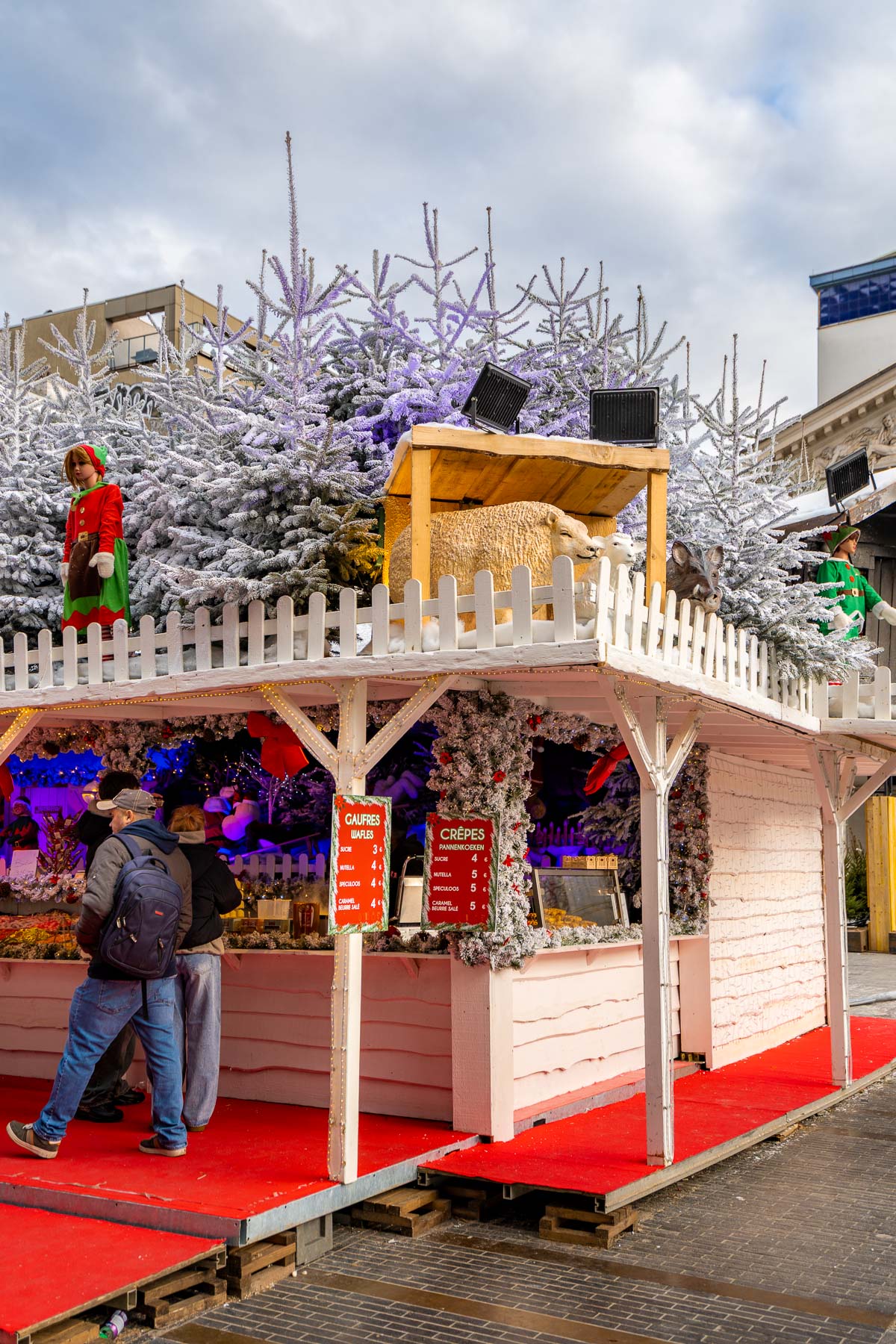 Festive food stall surrounded by snow-covered trees and Christmas decorations at Place de la Monnaie in Brussels, with red carpets and people ordering waffles and crêpes.