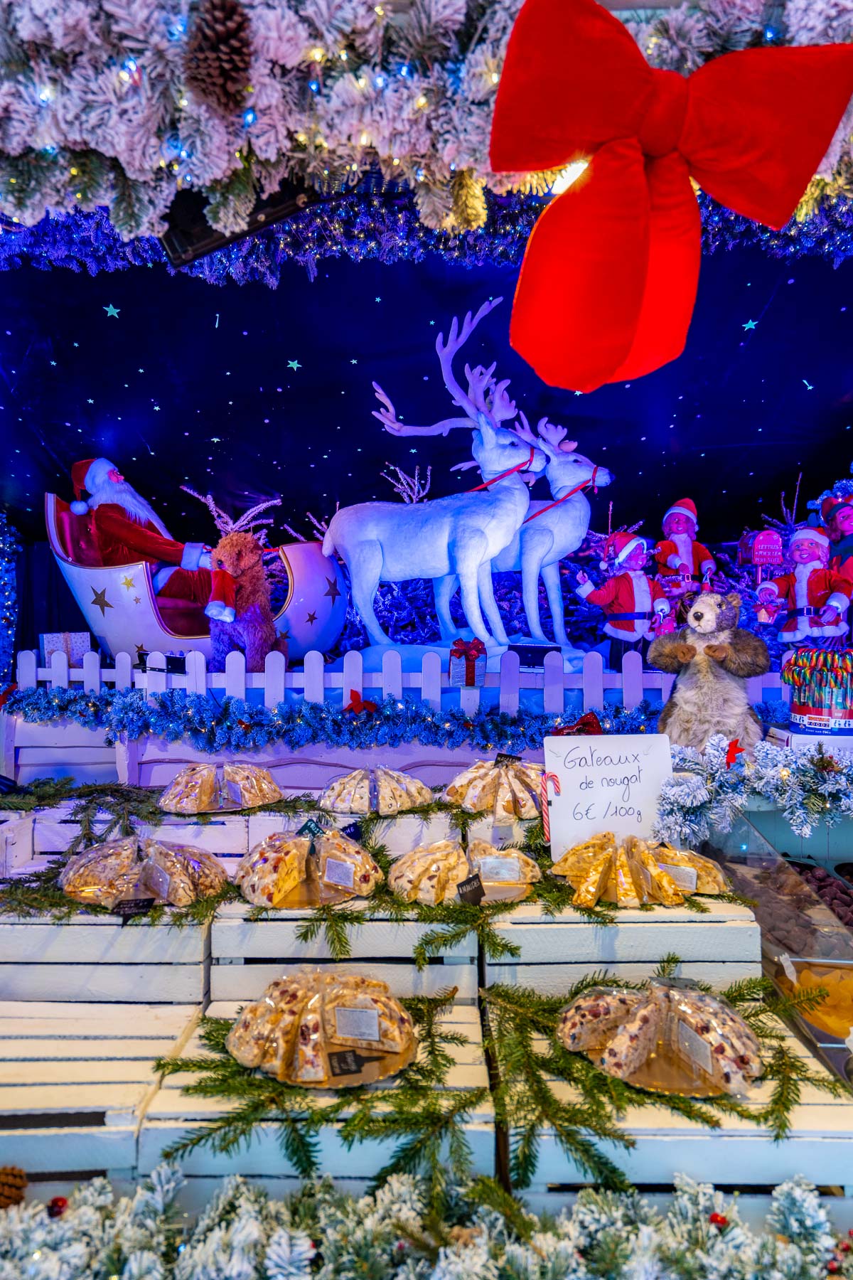 Close-up of a Christmas market stall in Brussels decorated with lights and festive figures, selling nougat and sweets with Santa and reindeer displays in the background.