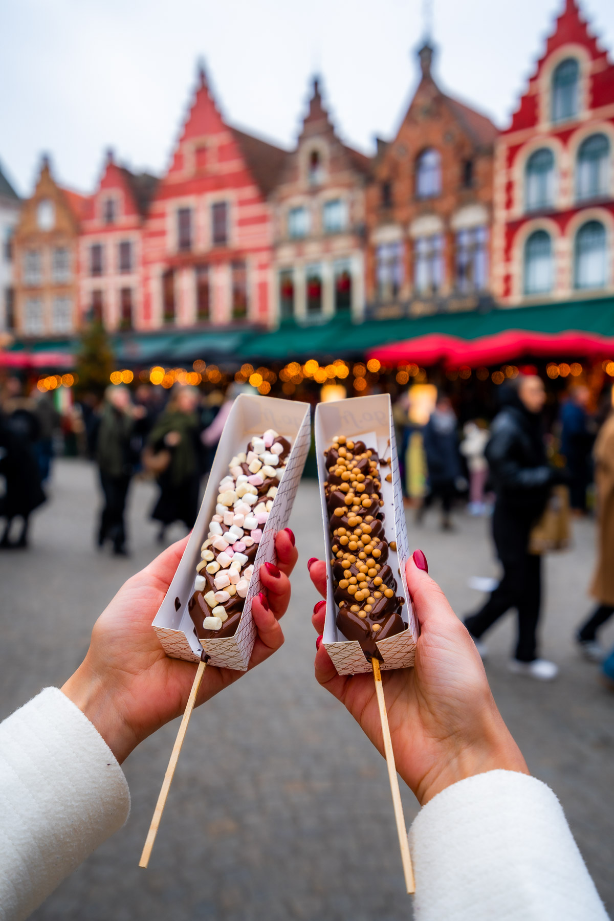 Close-up of two chocolate treats held up in front of the colorful gabled houses in Markt Square, Bruges, during the Christmas market.