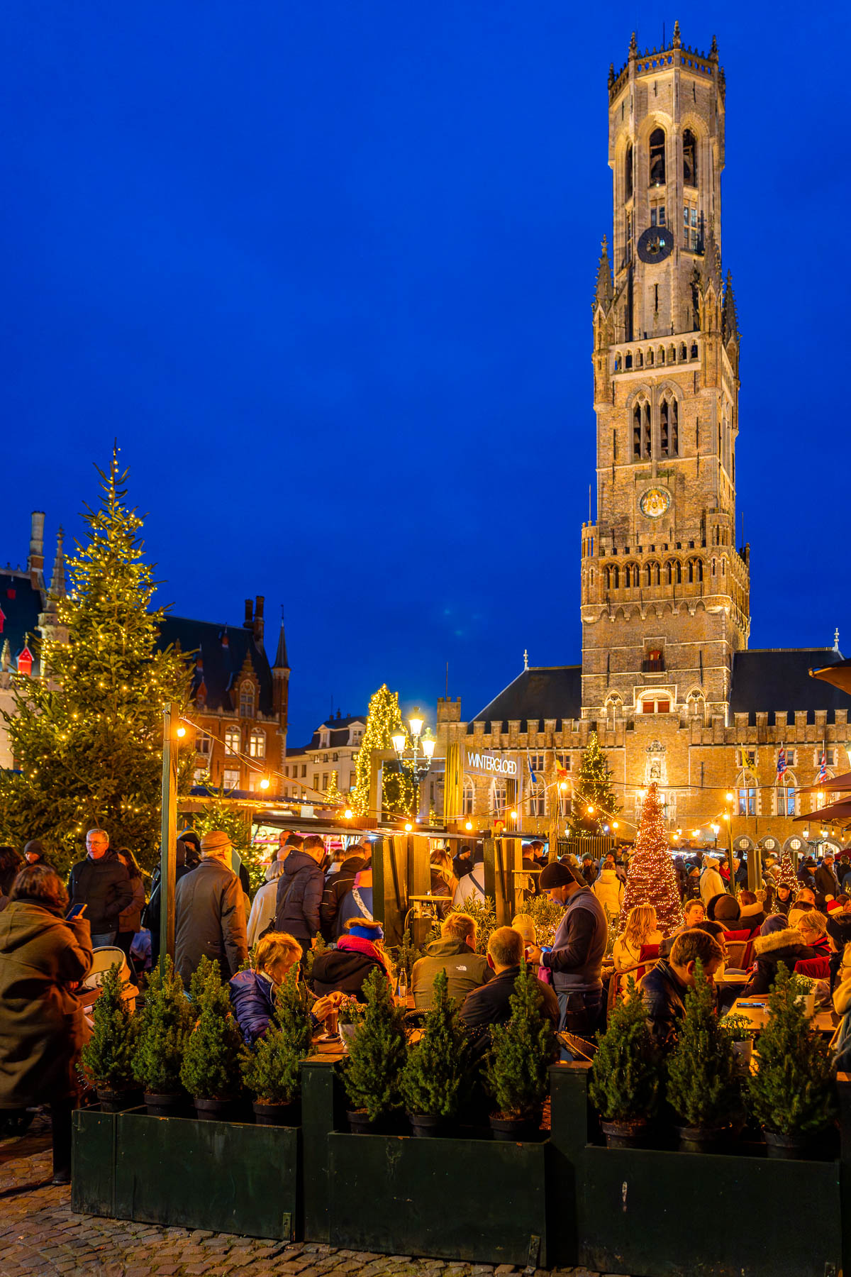 Crowds enjoying the Bruges Christmas market in Markt Square at night, with the Belfry of Bruges lit up in the background.