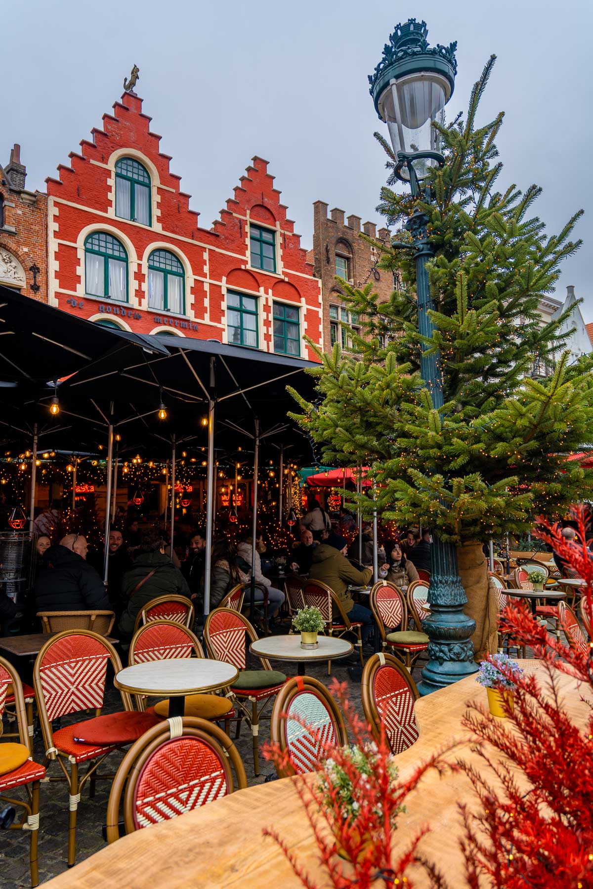Festive outdoor café in Bruges’ Markt Square with red chairs, Christmas trees, and people gathered under black umbrellas beside the colorful gabled buildings.