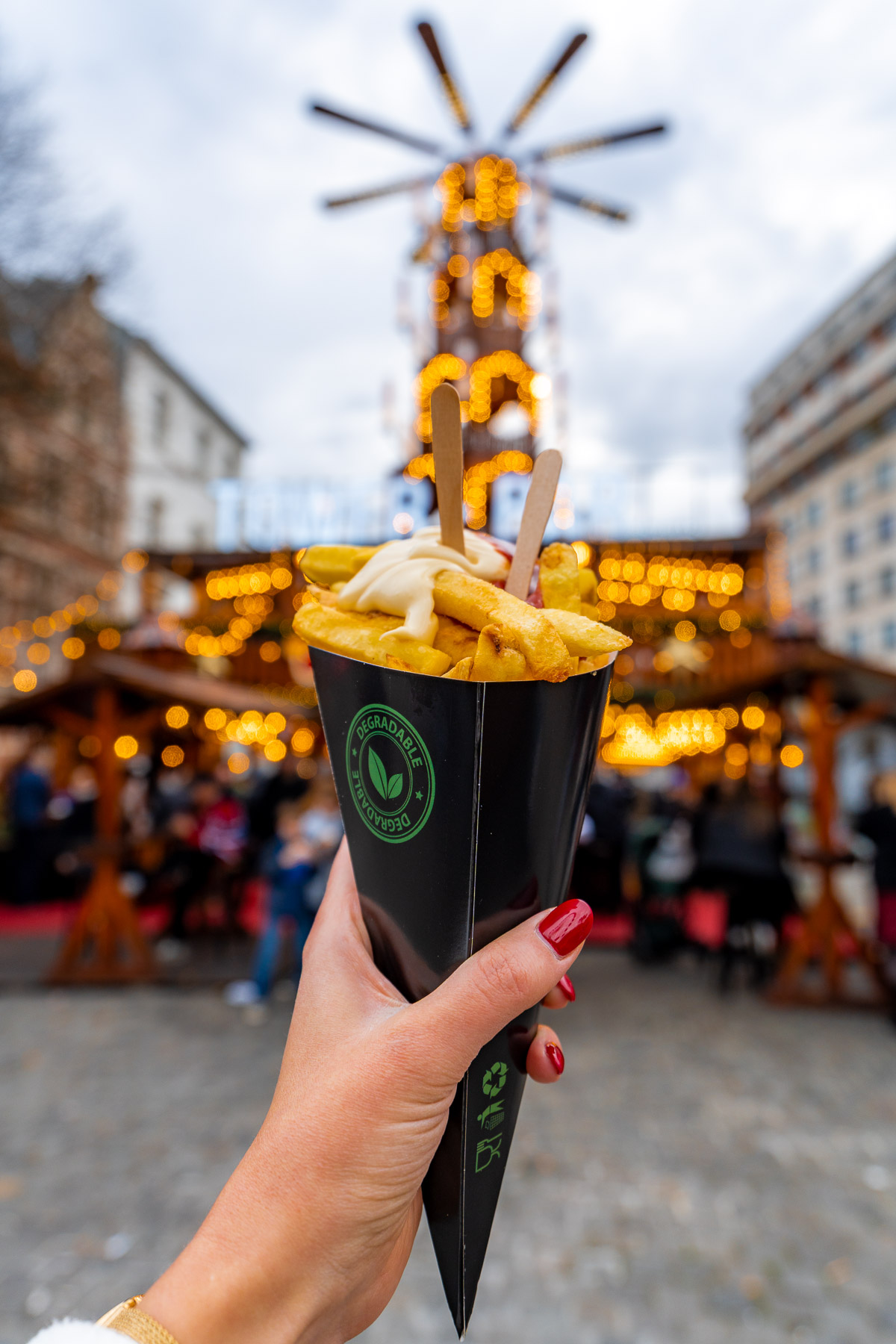Hand holding a cone of Belgian fries topped with mayo and ketchup at the Brussels Christmas market, with festive lights and a wooden tower blurred in the background.