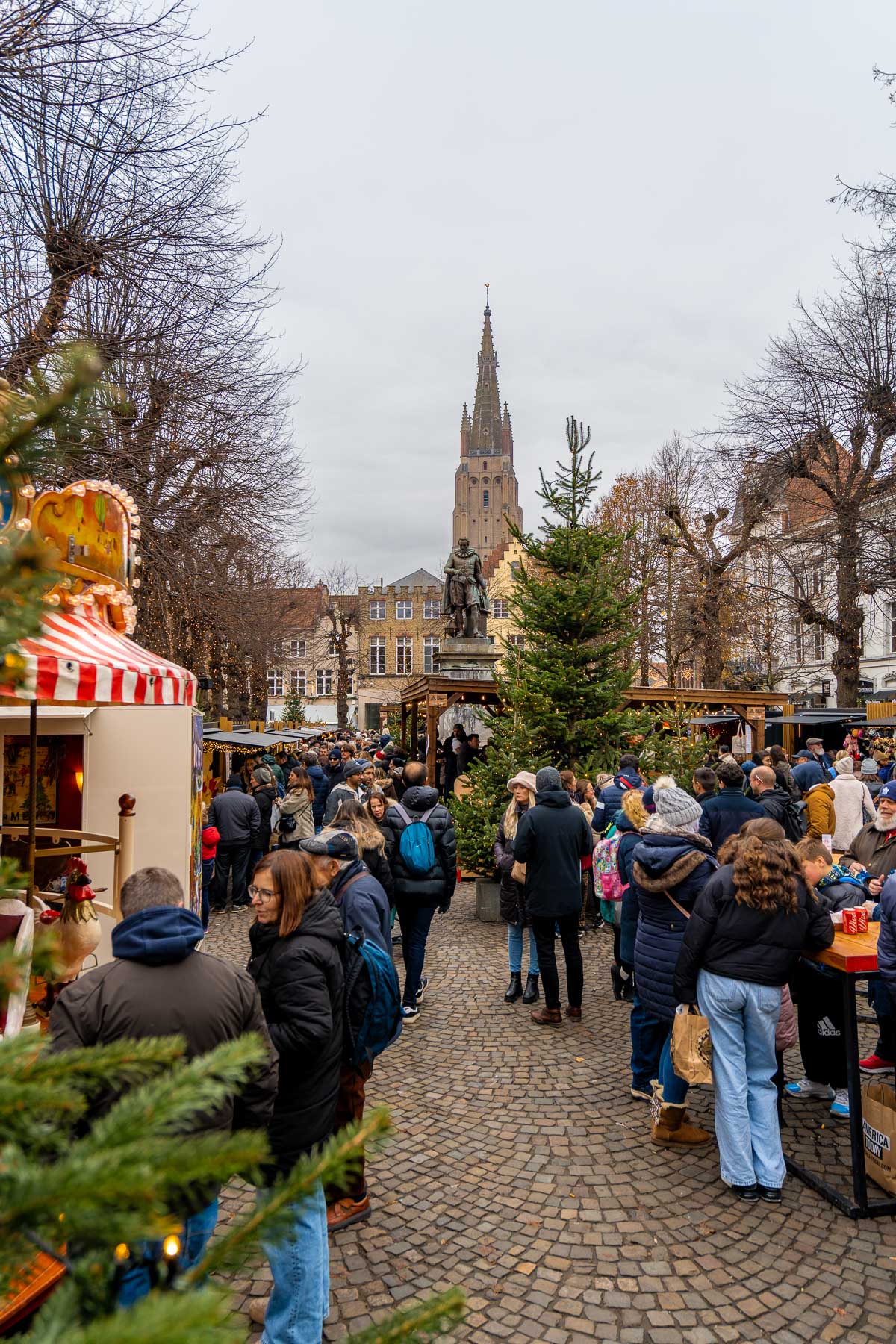 Crowds enjoying the Christmas market on Simon Stevinplein in Bruges, with festive trees and the Church of Our Lady visible in the background.