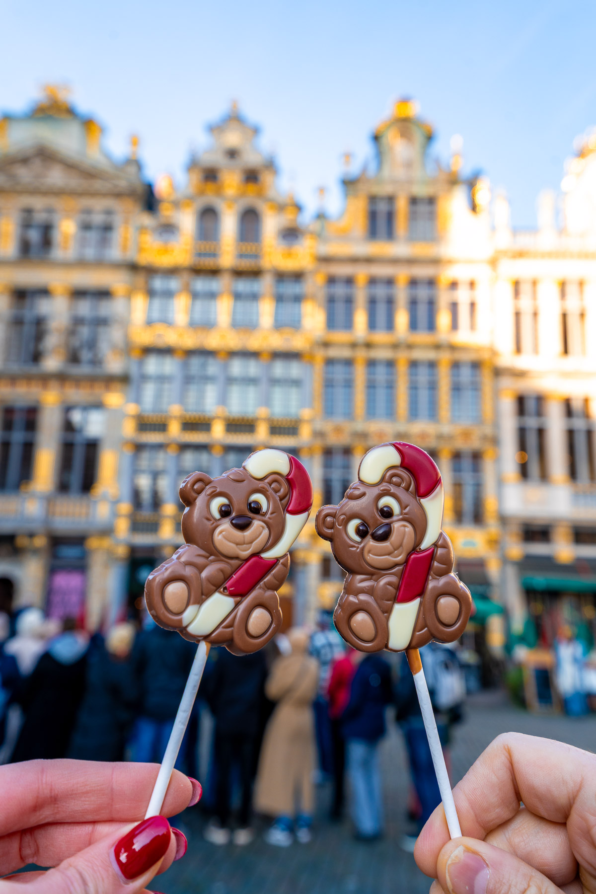 Two chocolate bear lollipops held up in front of the ornate golden buildings of Grand Place in Brussels on a sunny day.