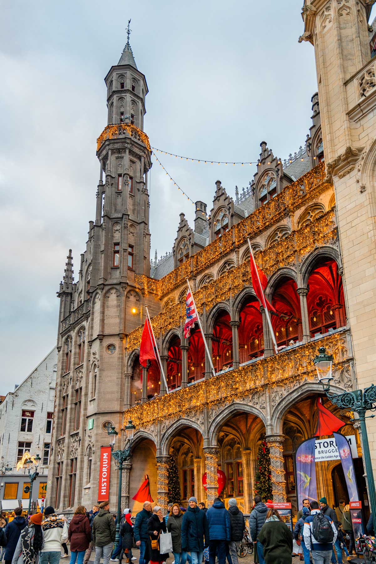 The illuminated facade of Historium Brugge decorated with Christmas lights, as people walk around the festive square below.