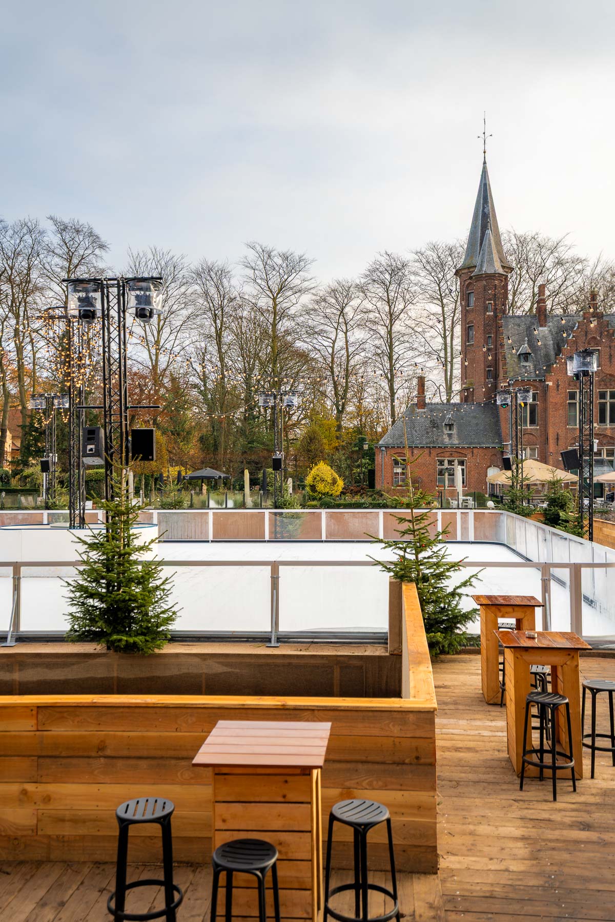Outdoor ice rink at Minnewaterpark in Bruges, surrounded by small pine trees and wooden seating areas, with a historic brick building and tower in the background.