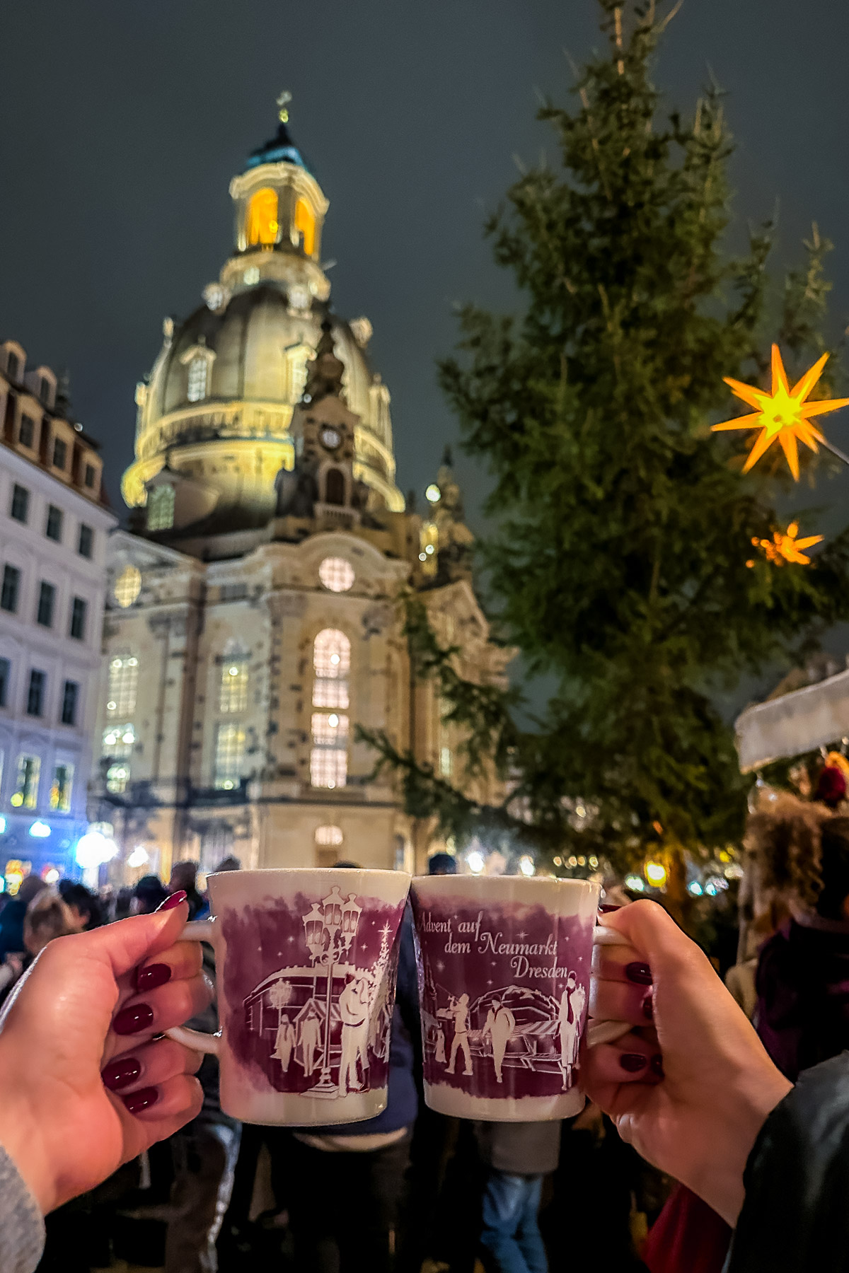 Two hands holding festive mugs of mulled wine at the Neumarkt Christmas Market in Dresden, with the illuminated Frauenkirche in the background.