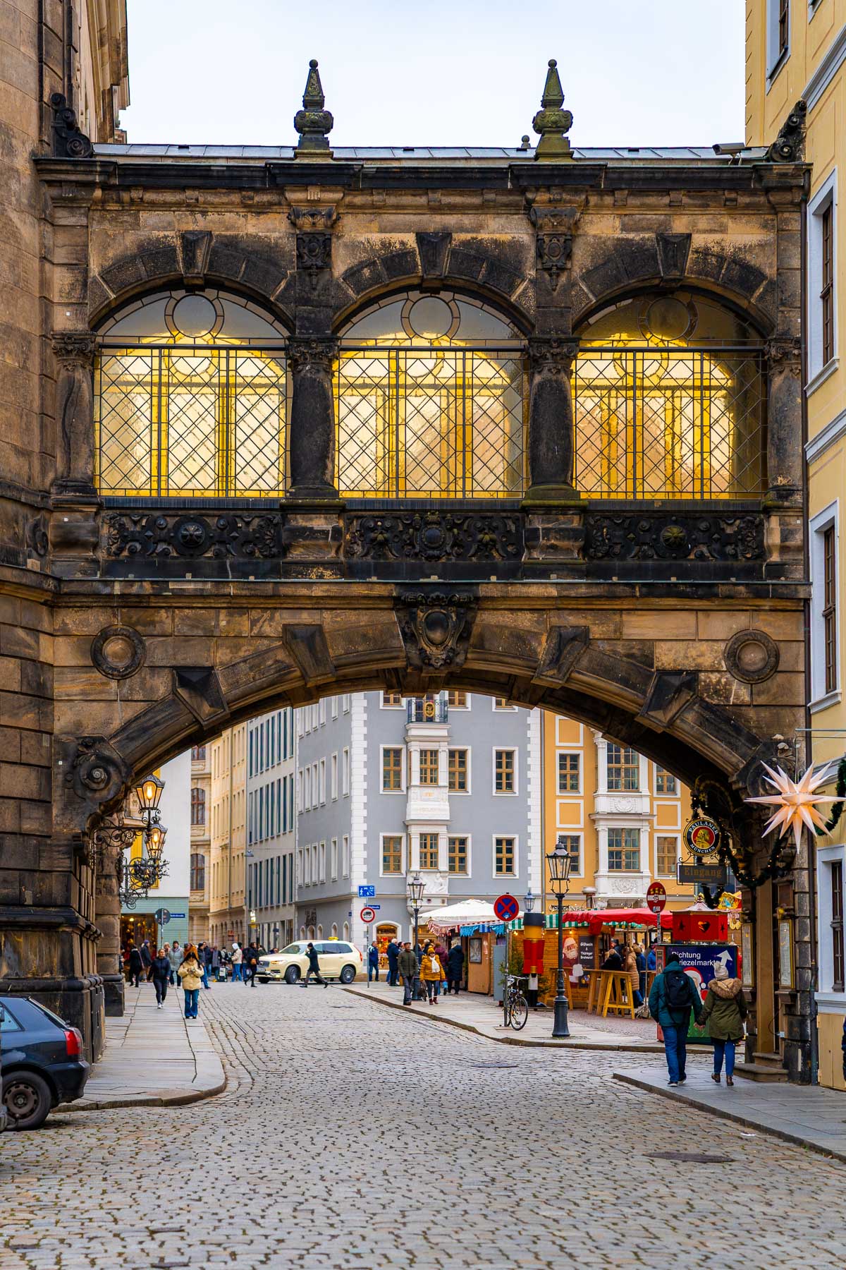 People walking through the cobblestone streets of Dresden’s Old Town, passing under the illuminated bridge of the Stallhof during the Christmas market season.