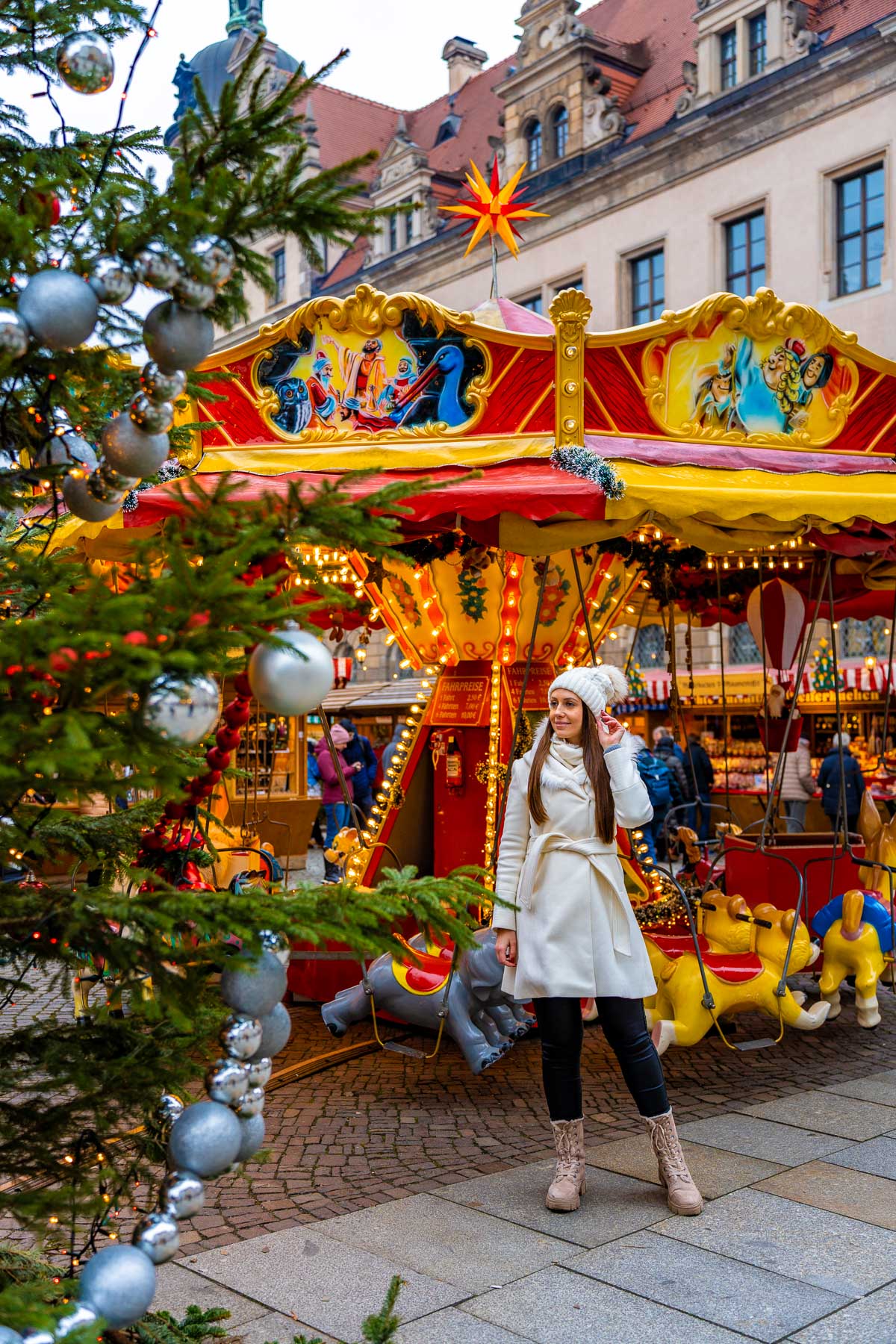 A woman dressed in a white coat standing near a carousel at Dresden’s Romantic Christmas Market, surrounded by Christmas trees and festive lights.