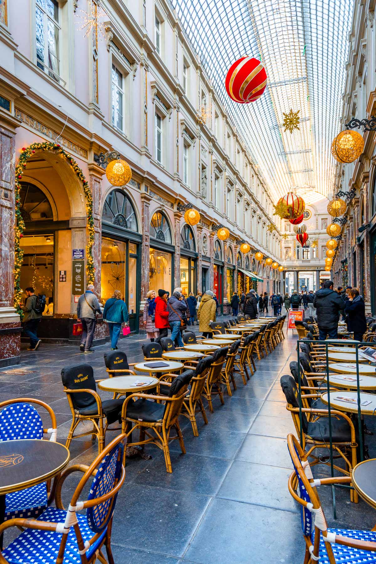 Shoppers and café tables inside the Royal Gallery of Saint Hubert in Brussels, decorated with oversized red and gold ornaments for the holiday season.