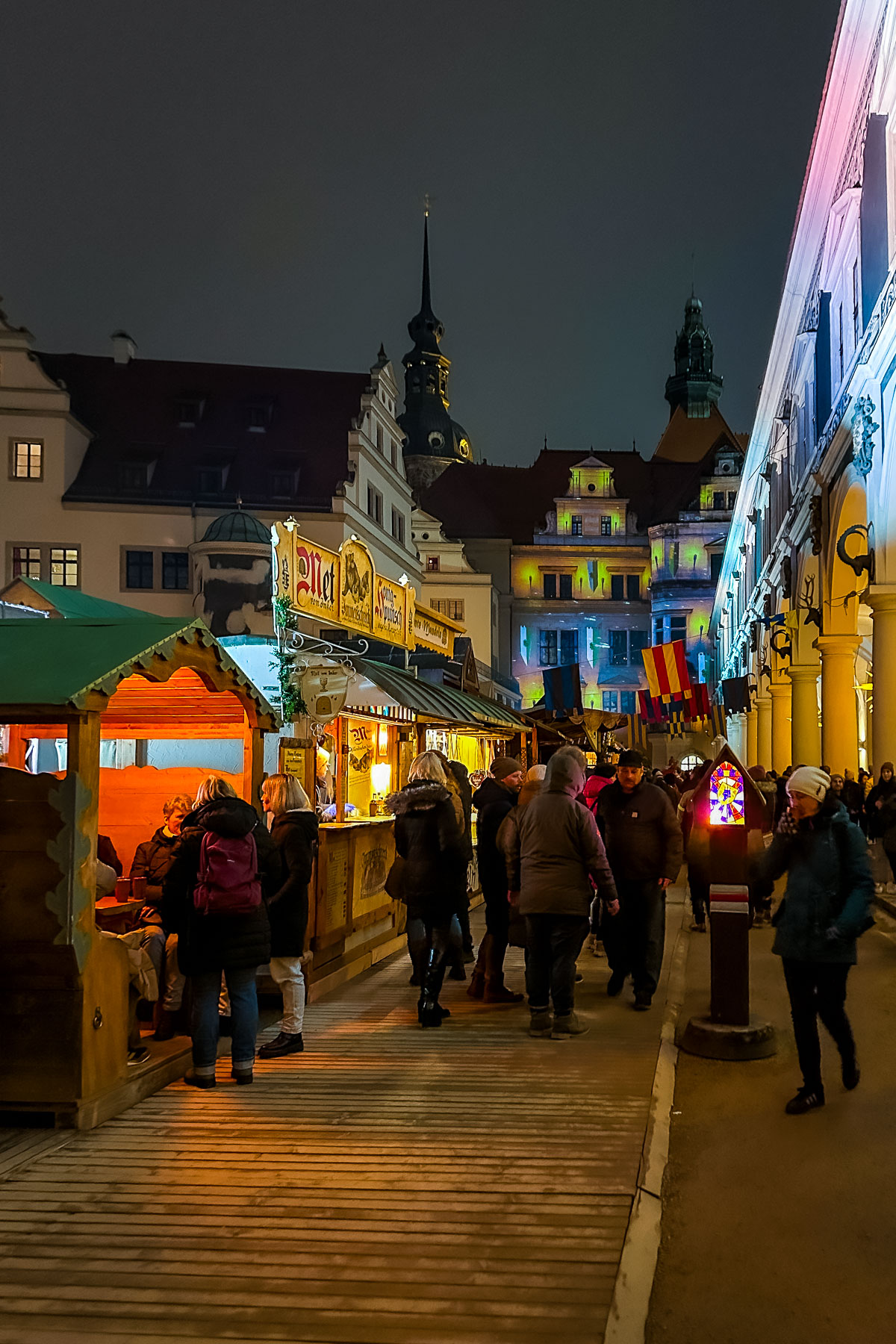 Visitors walking through the Stallhof Medieval Christmas Market in Dresden at night, surrounded by wooden stalls and colorful lights against the historic buildings.