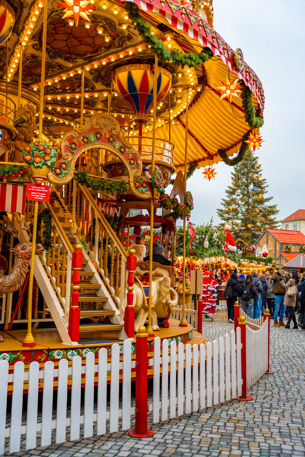 A brightly lit vintage carousel decorated with garlands and stars at the Striezelmarkt in Dresden, with visitors walking nearby.
