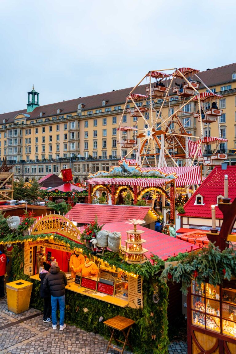 View of the Striezelmarkt in Dresden showing the Ferris wheel, festive stalls, and Christmas decorations in front of the city’s historic buildings.