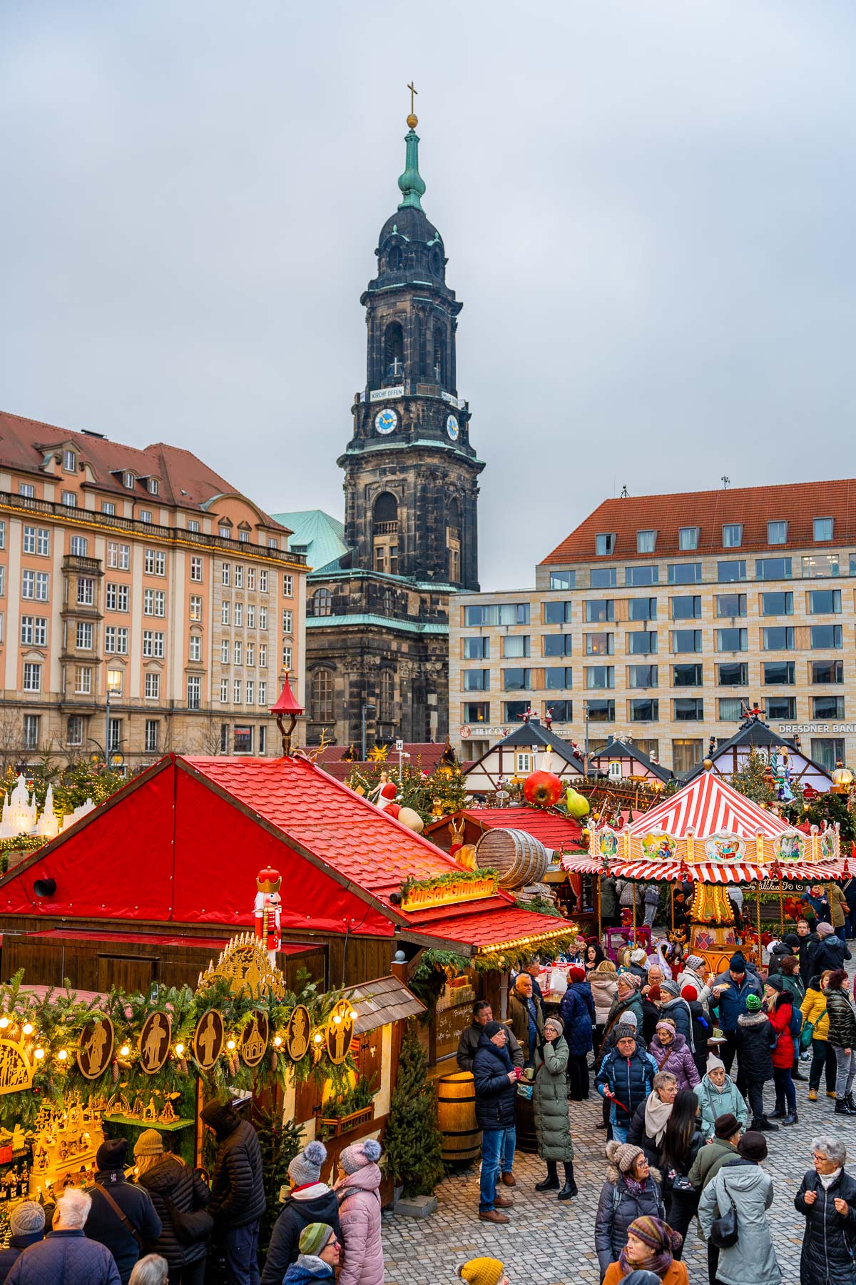 Daytime view of the Striezelmarkt in Dresden with festive stalls, a carousel, and the black church tower rising in the background.