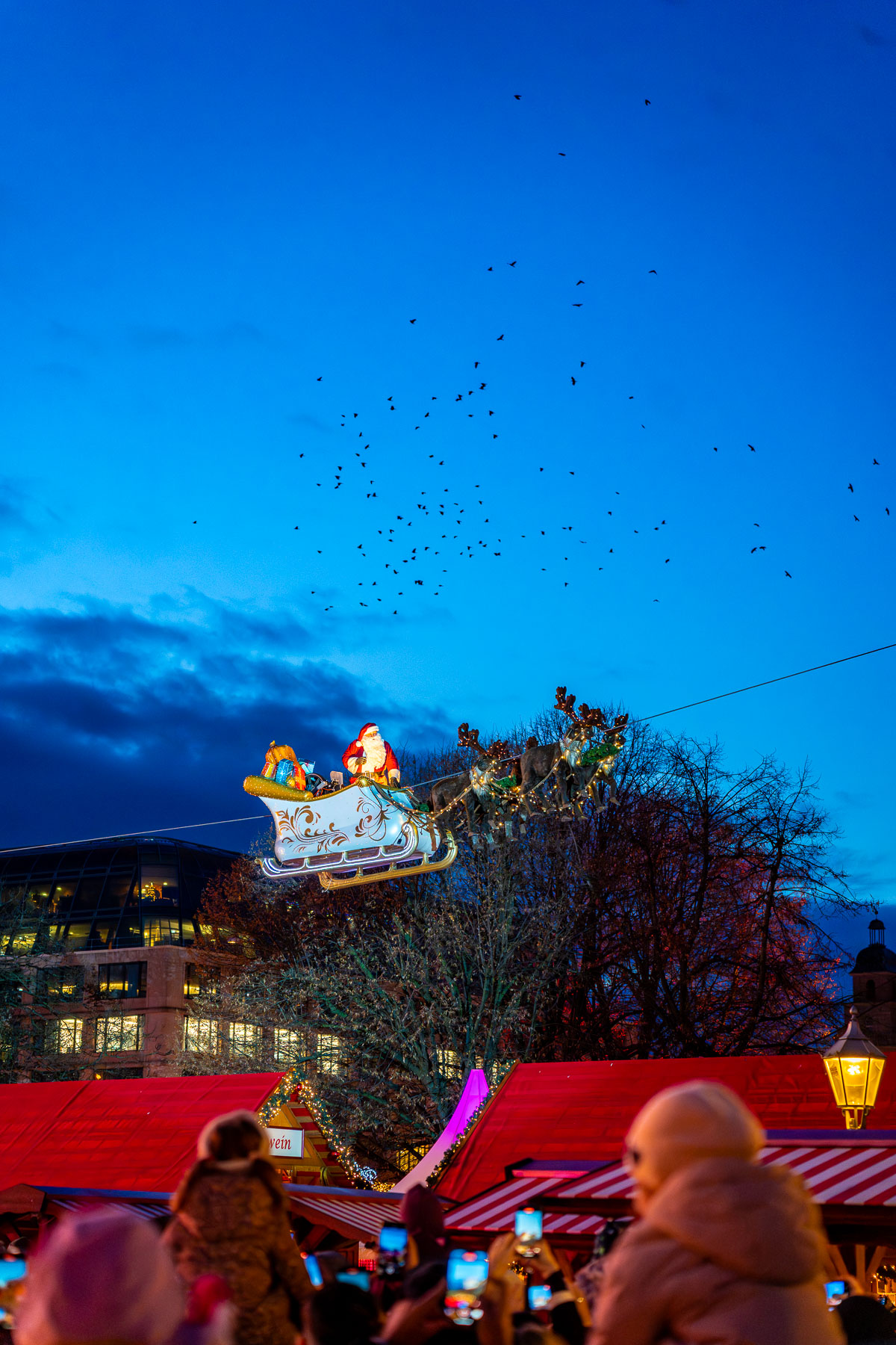 Santa Claus decoration flying in a sleigh above the Christmas market at Rotes Rathaus in Berlin, with red-roofed stalls below and people in the foreground holding up phones.