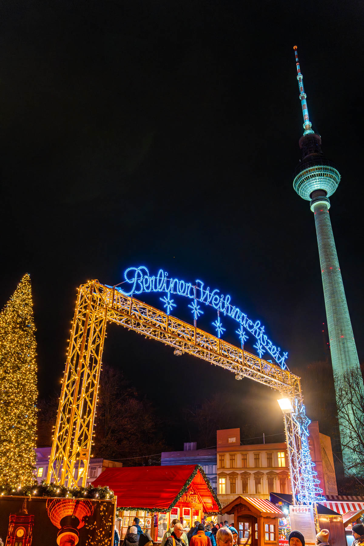 The entrance arch of Berliner Weihnachtszeit at Rotes Rathaus in Berlin at night, with illuminated lettering, Christmas stalls underneath, and the TV Tower visible in the background.