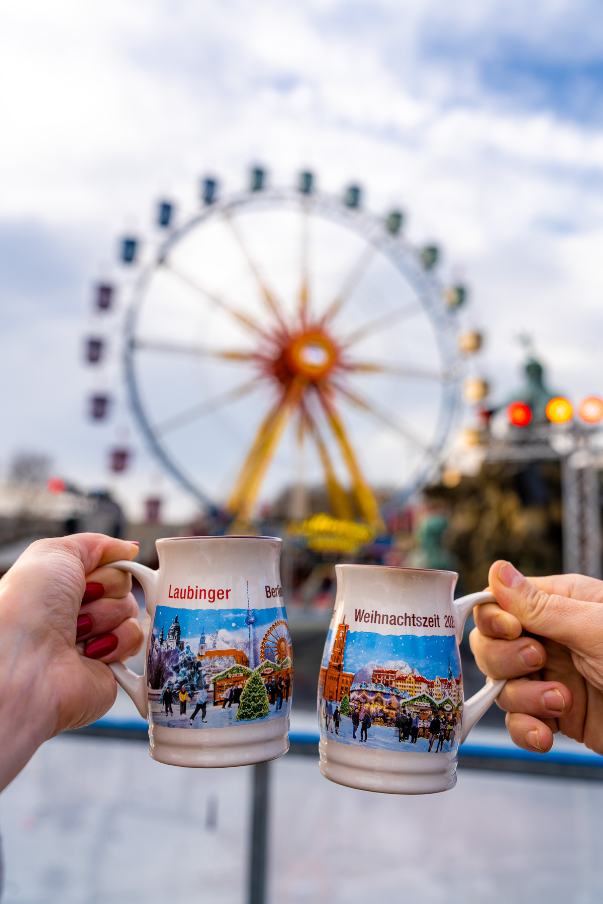 Two hands holding Berlin Christmas market mugs in front of the Ferris wheel at Berliner Weihnachtszeit at Rotes Rathaus, with the wheel visible in the background.