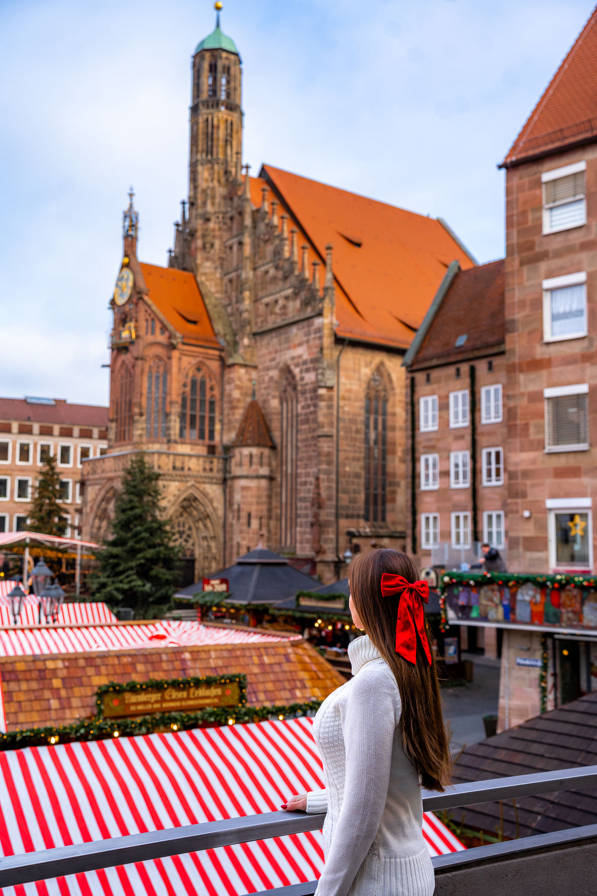 A woman with a red bow in her hair stands on a balcony overlooking the Christkindlesmarkt in Nuremberg, with red and white striped market stalls below and the Church of Our Lady in the background.