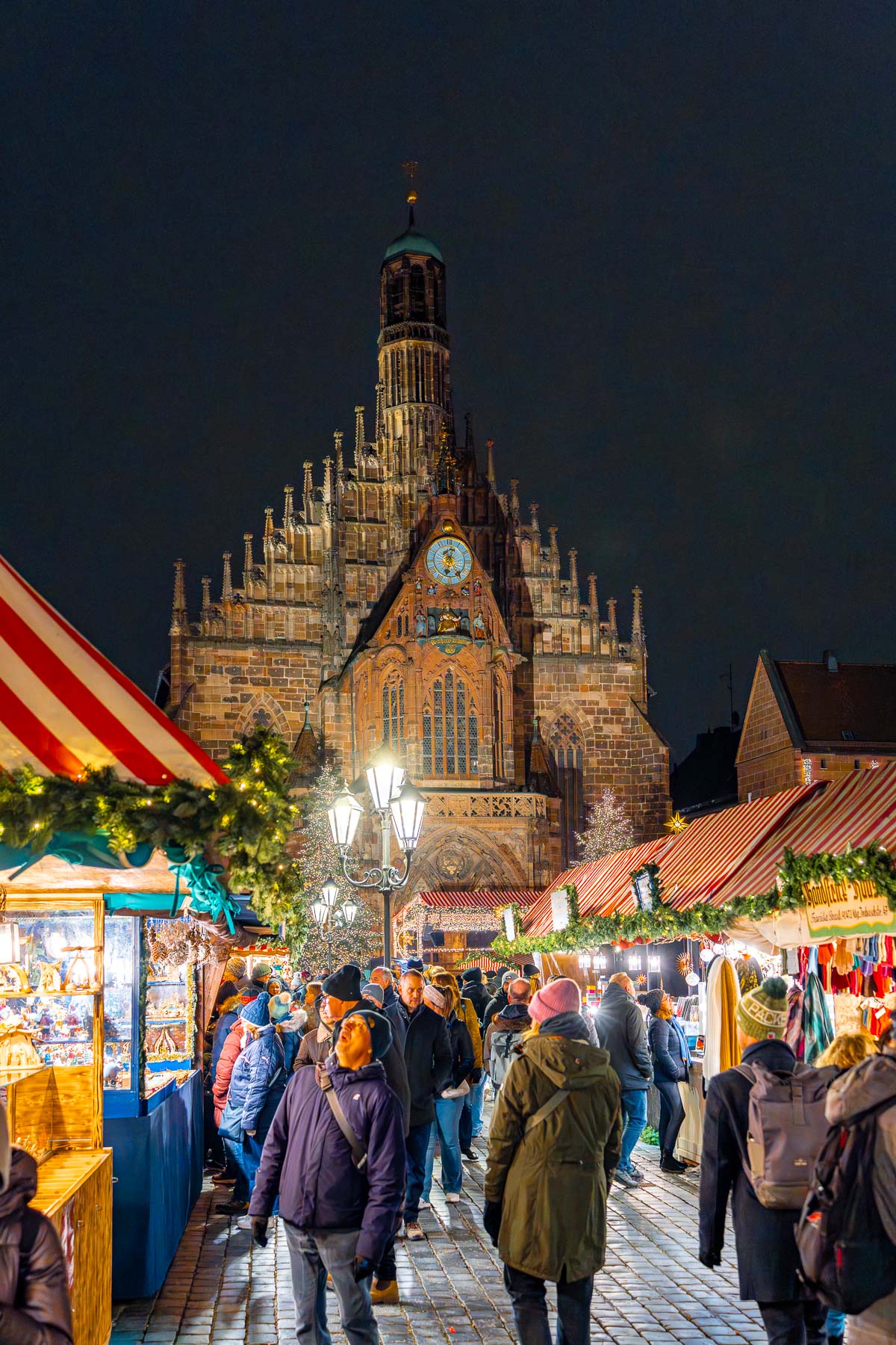 Crowds walking through the Christkindlesmarkt in Nuremberg at night, with illuminated wooden stalls on both sides and the Church of Our Lady rising above the market.