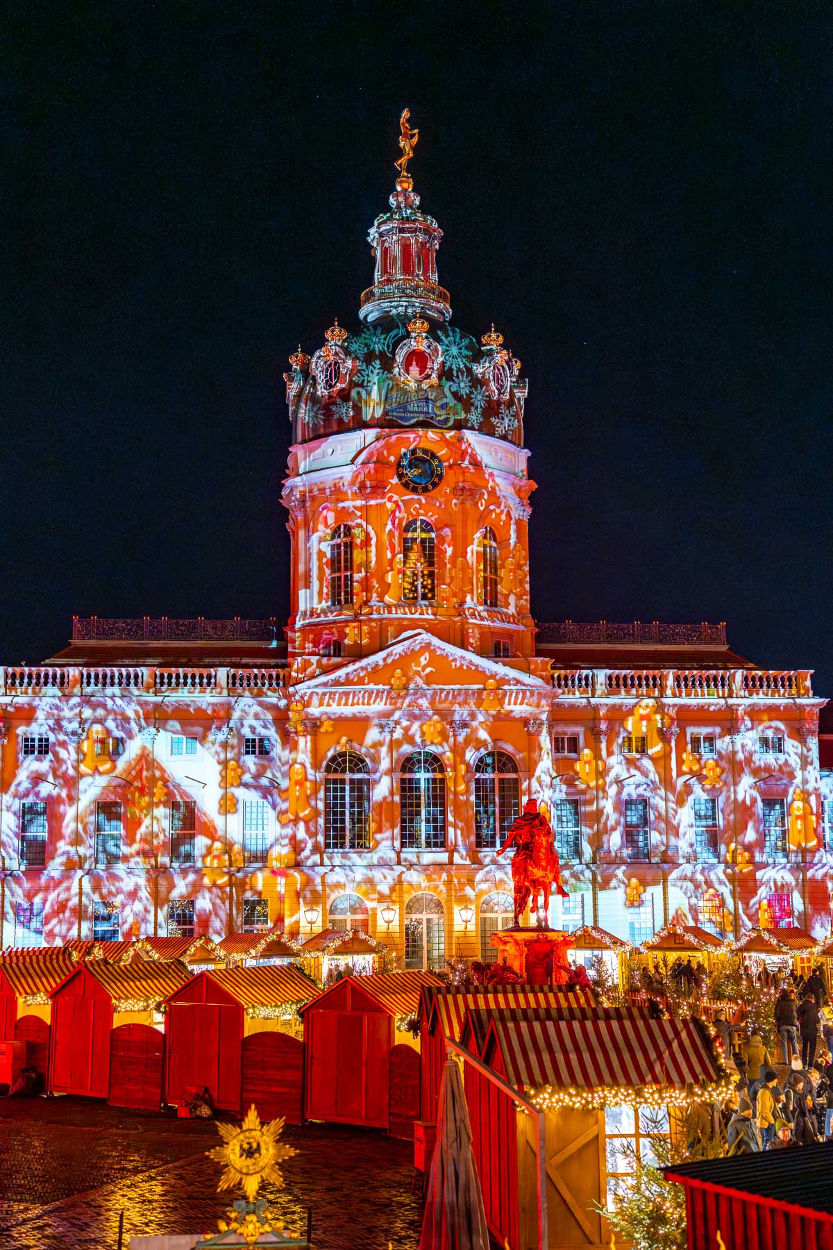 Charlottenburg Palace in Berlin is lit with projection lights behind rows of red Christmas market stalls and a statue in the foreground.