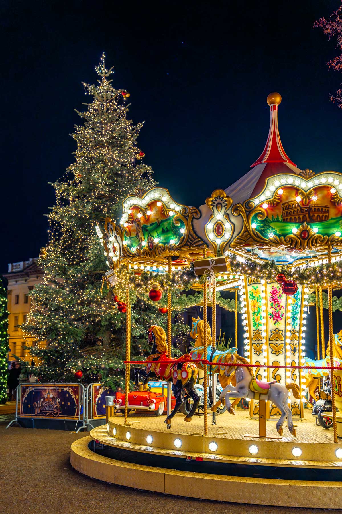 A carousel with painted horses and lights stands next to a large Christmas tree at the Christmas Market at Charlottenburg Palace, Berlin, with market stalls and historic buildings in the background at night.